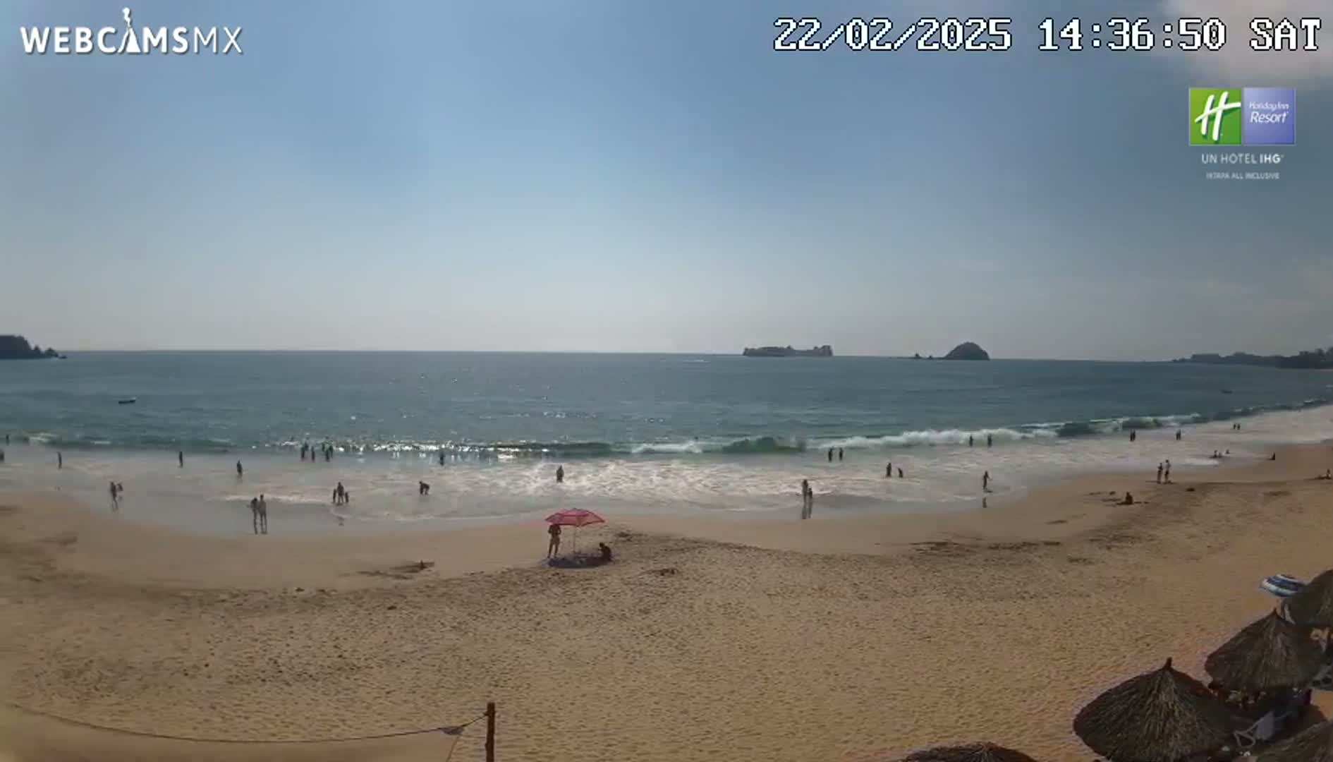 A sandy beach with people swimming and relaxing under a mostly sunny sky, with small waves breaking on the shore and islands visible in the distance.