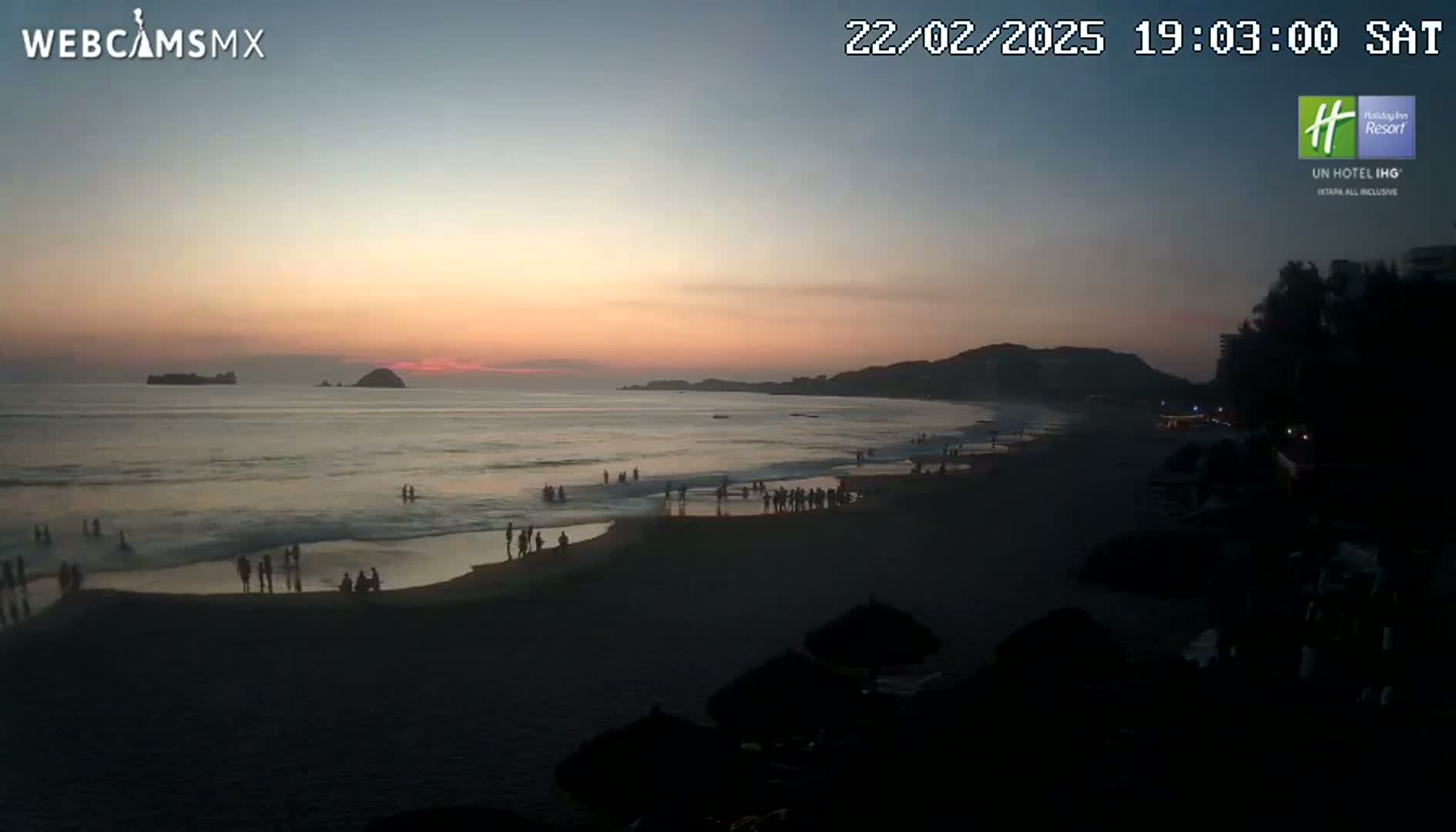 A beach scene at sunset shows people on the sand and in the shallow water, with calm ocean waves and a dark silhouette of land in the background.