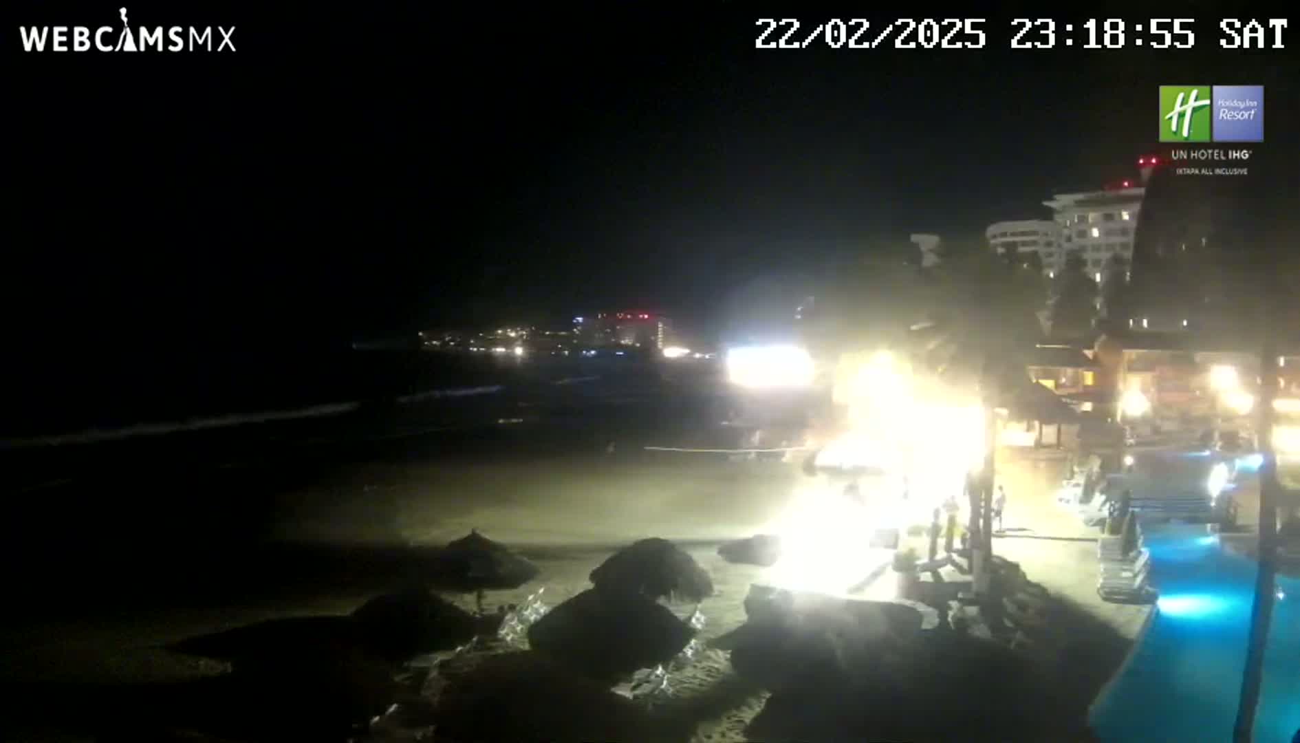 A nighttime beach scene shows a bright light emanating from a beachfront resort near a pool, with dark ocean waves lapping the shore under a clear night sky.
