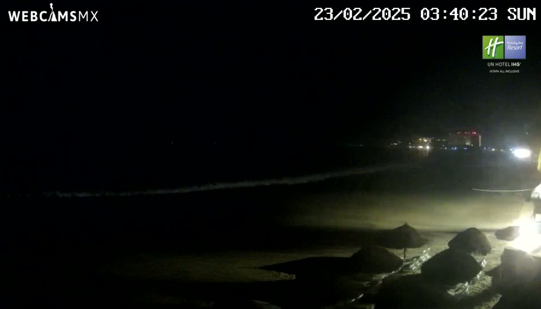 A dark nighttime beach scene shows a dimly lit shoreline with several closed beach umbrellas under a clear, starless sky.