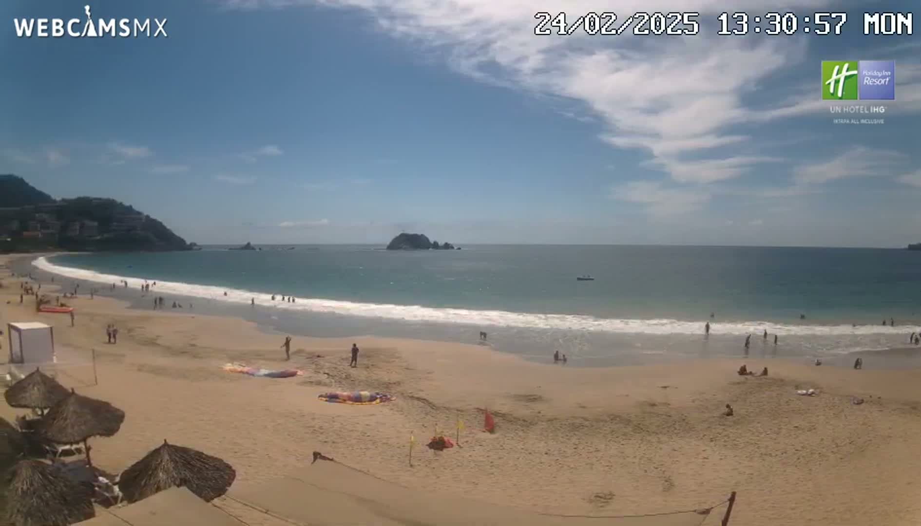 A mostly sunny day at a sandy beach with calm waves, people swimming and relaxing, and a mountain in the background.