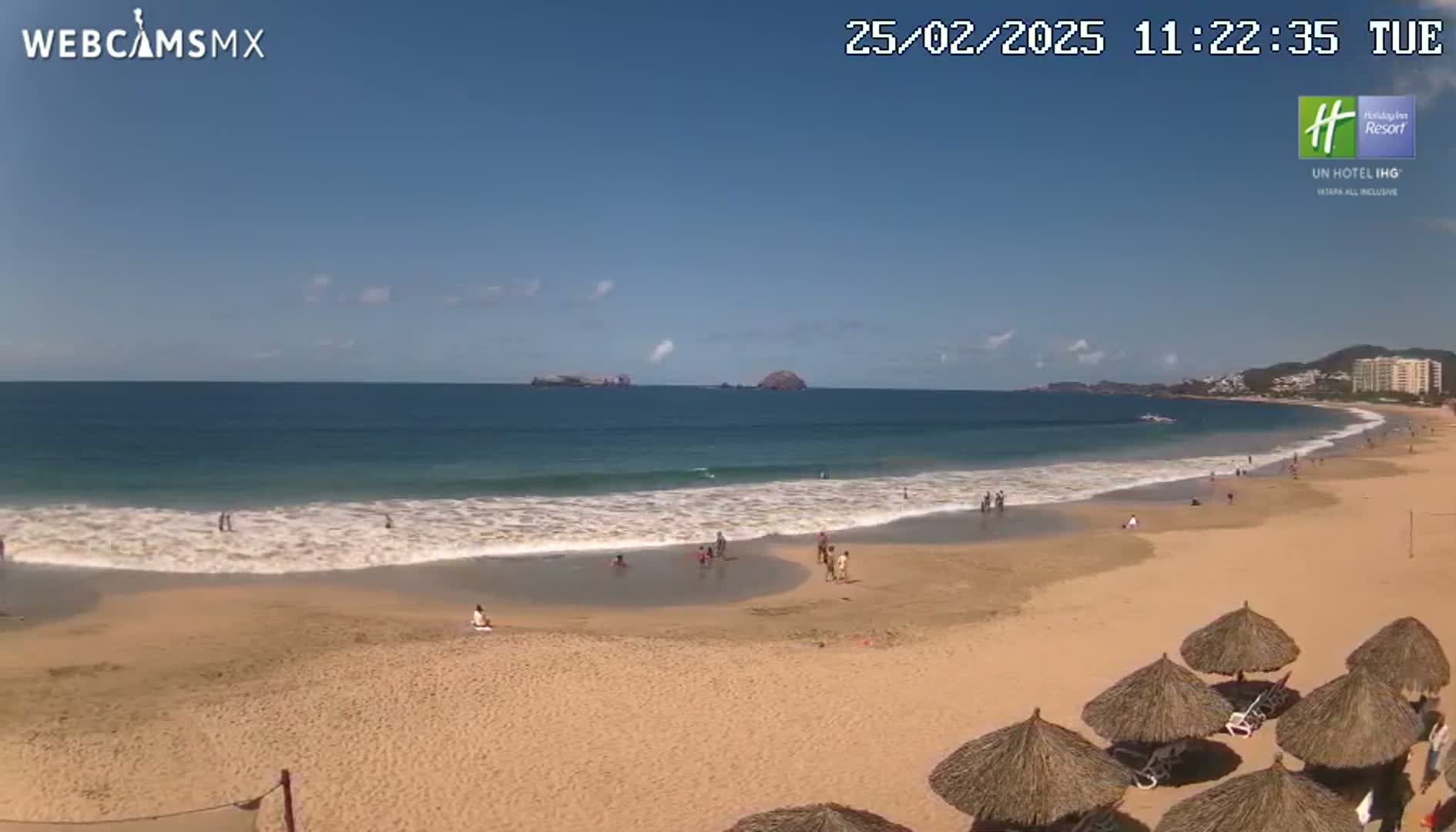 A sandy beach with calm waves and a few people swimming and sunbathing under a mostly clear sky.