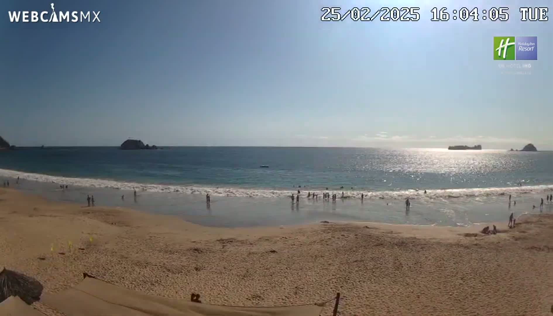 A sandy beach with calm waves and a few people on it stretches out to meet a calm ocean under a sunny sky.