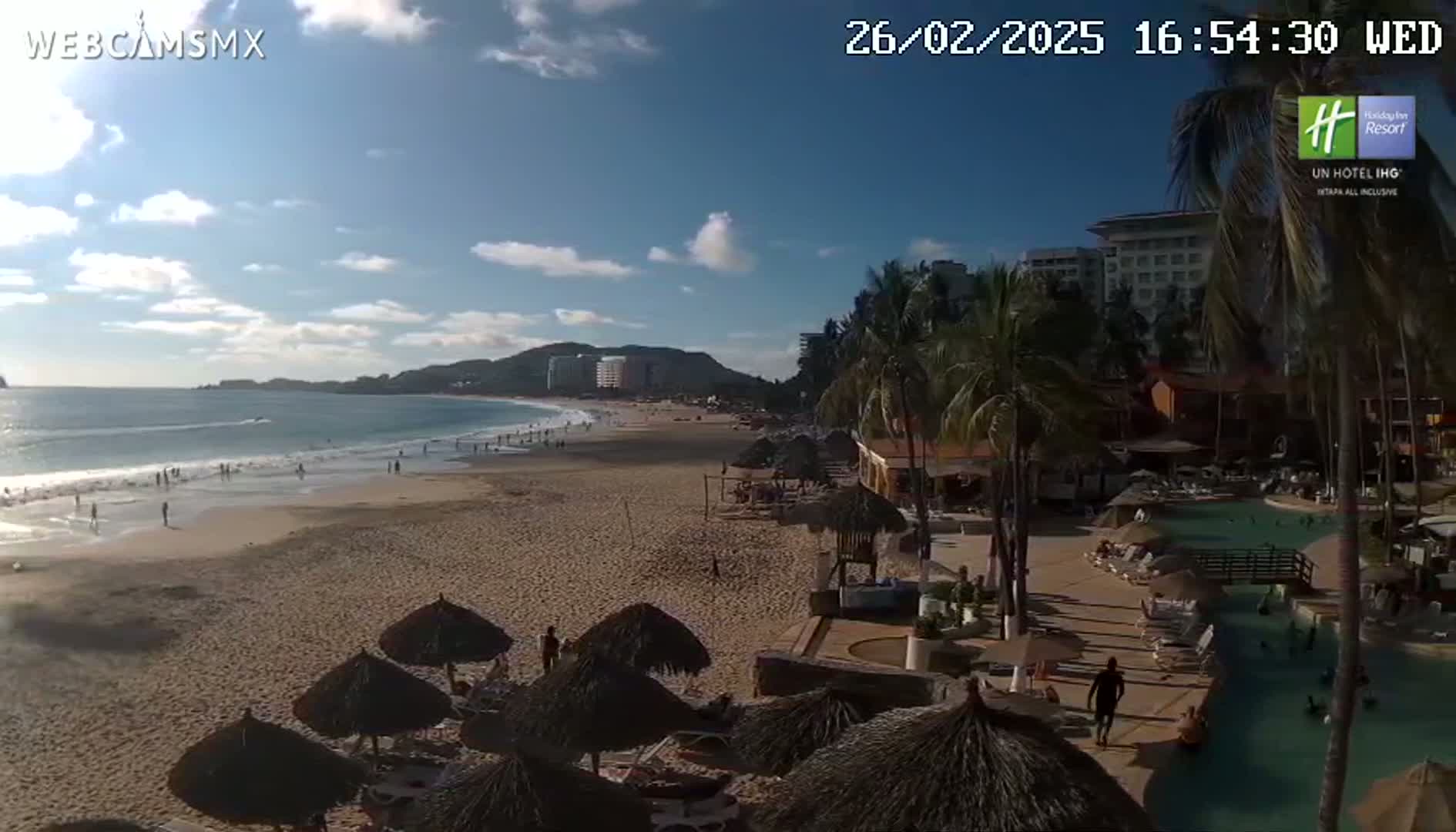 A sunny beach with calm waves, palm trees, beach umbrellas, and a resort with a pool is visible under a mostly clear sky.