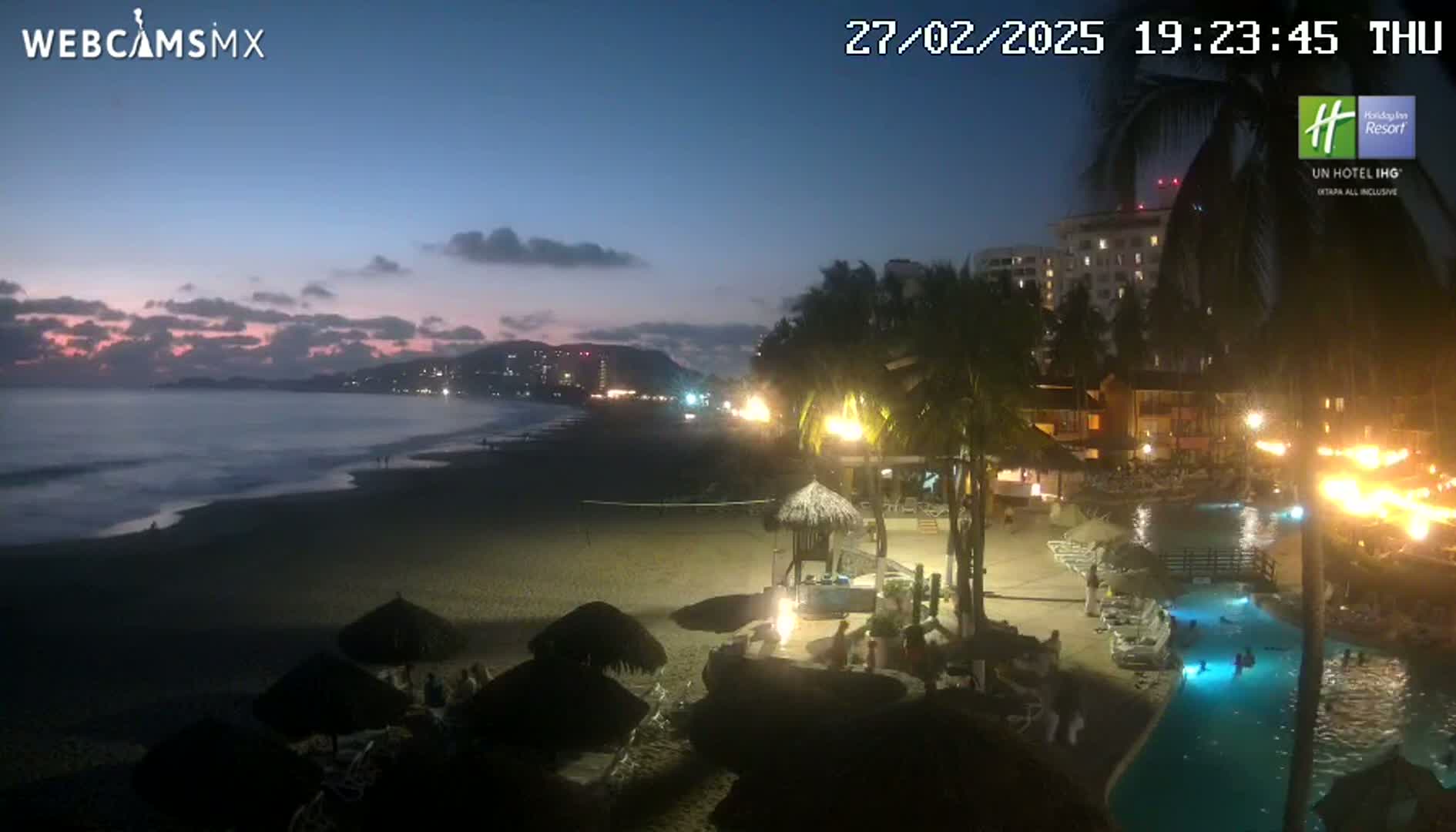 A nighttime beach scene shows a dark ocean with waves, a sandy shoreline with beach umbrellas and people, a resort with lights and a swimming pool, all under a twilight sky.