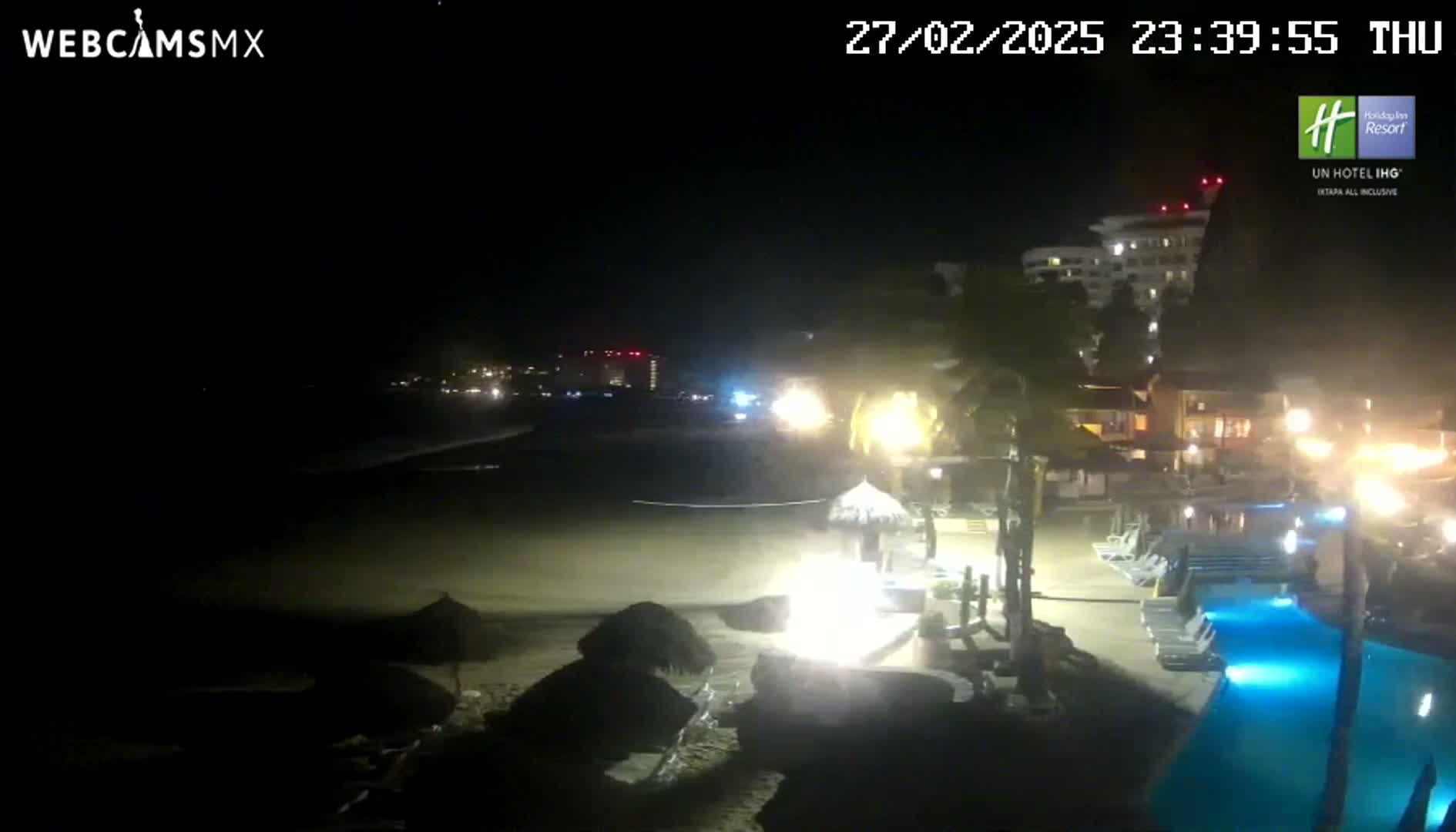 A nighttime view of a beach resort with a lit-up pool and palm trees, under a clear, dark sky.