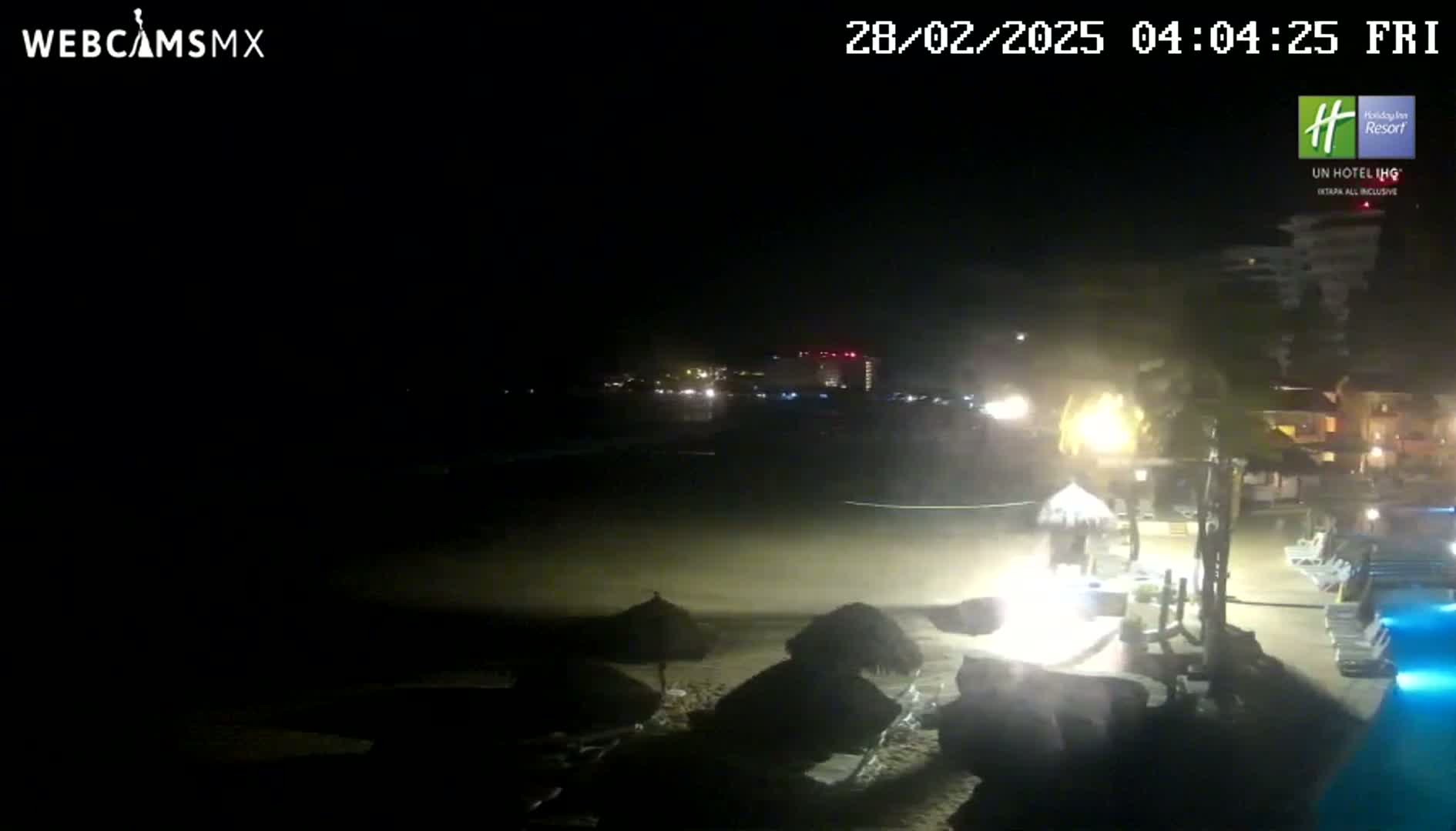 A nighttime beach scene shows a dark sandy beach with a few closed umbrellas, illuminated buildings in the background, and palm trees swaying in the wind.