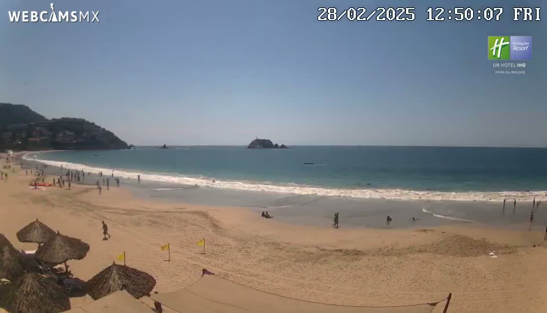 A sandy beach with several people and beach umbrellas stretches along a calm, blue ocean under a clear, sunny sky, backed by hills and a small island in the distance.