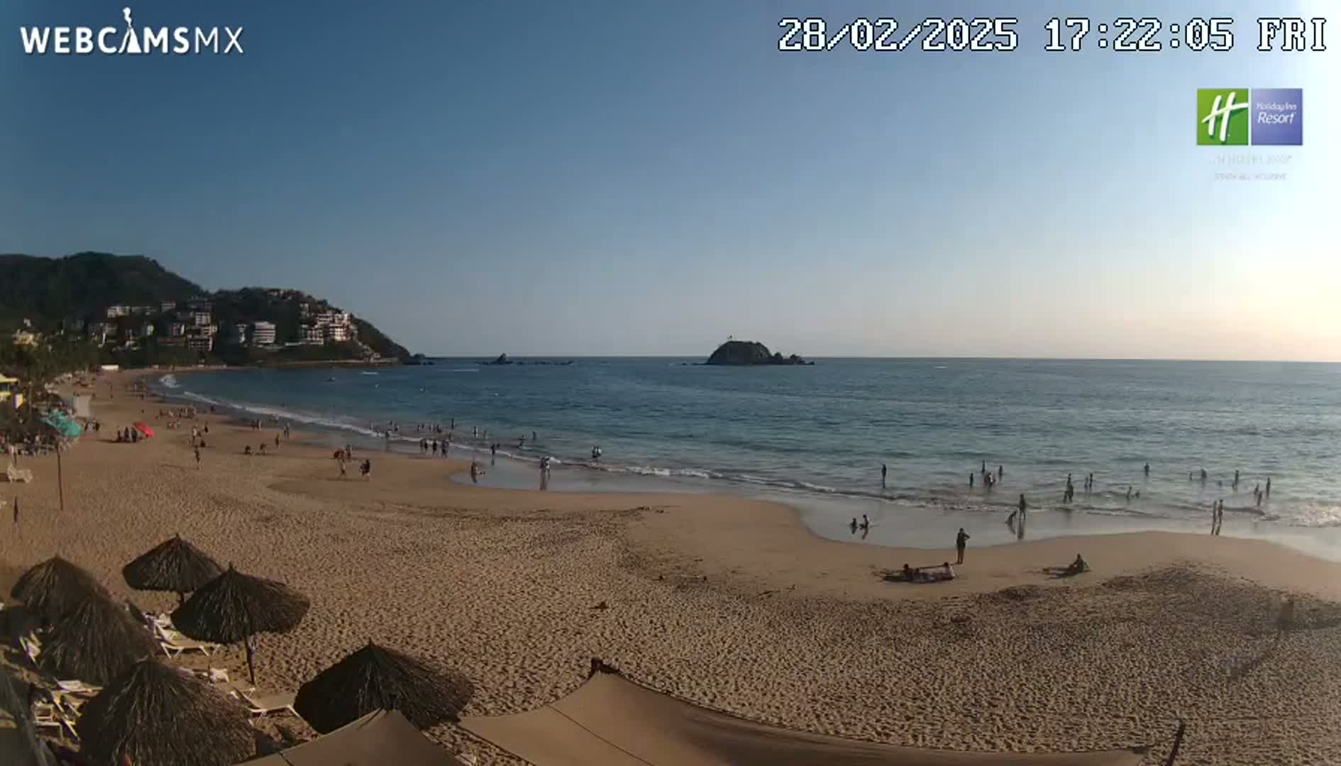 A sandy beach with a calm ocean and a few people swimming and sunbathing under a clear blue sky.