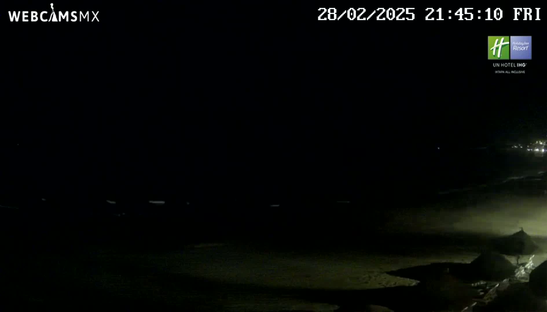 A dark nighttime beach scene shows a mostly-black ocean with a few faint lights in the distance and dark shapes on the shore.