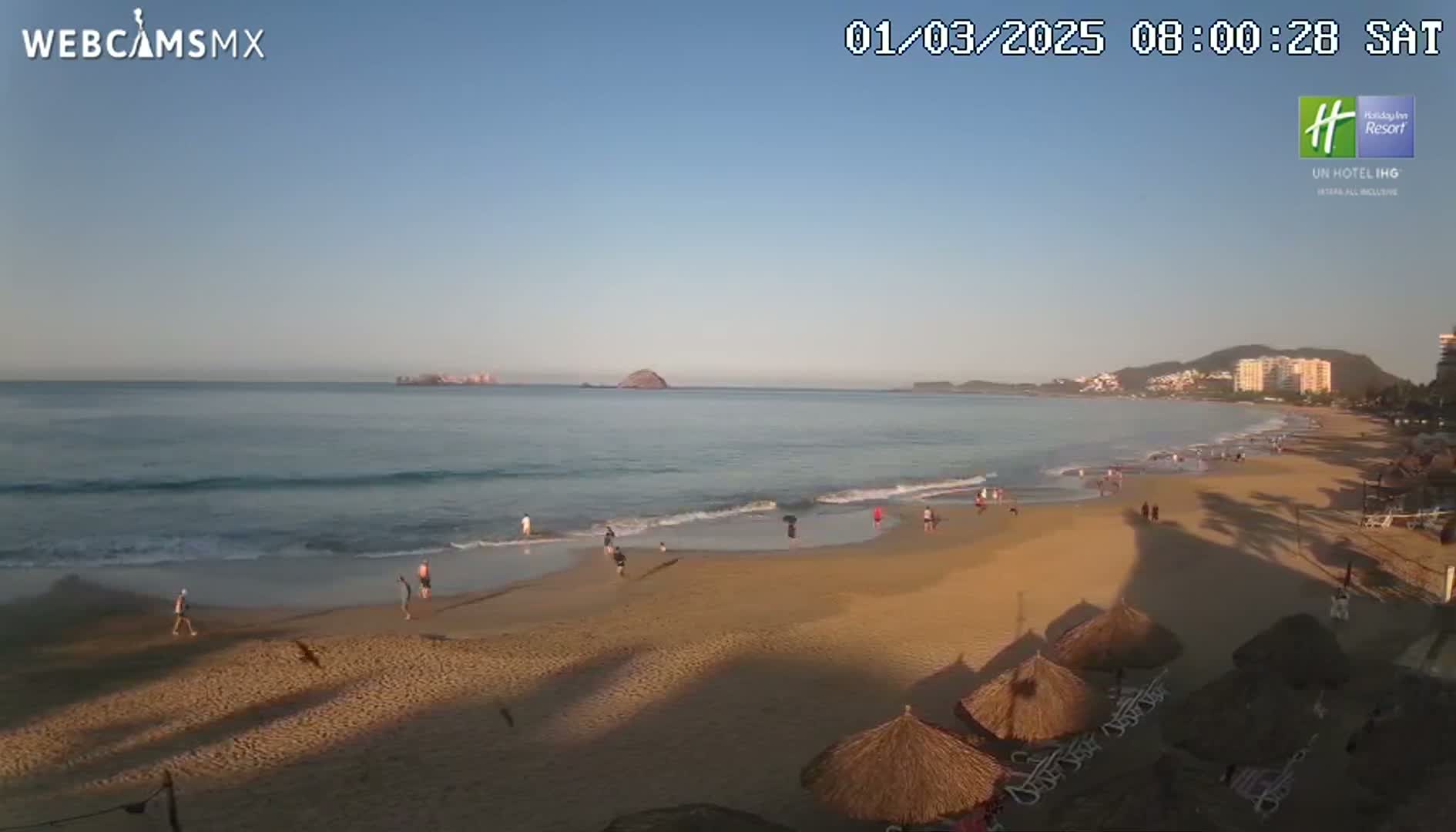 A mostly sunny beach scene shows a calm ocean with small waves lapping a sandy shore where several people stroll, with thatch umbrellas and lounge chairs visible in the foreground.