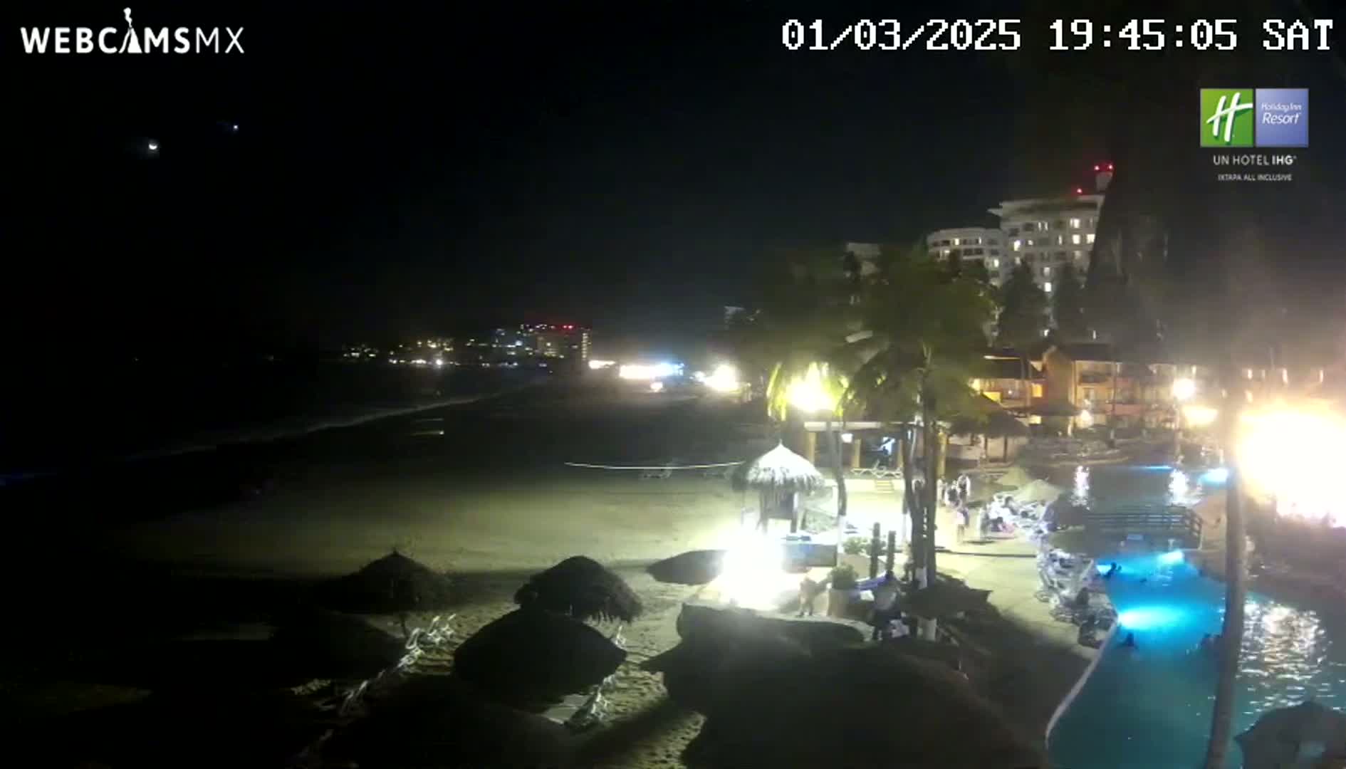 A nighttime beach scene shows a sandy shoreline with palm trees, resort buildings, and a swimming pool lit up with blue lights under a clear, dark sky.