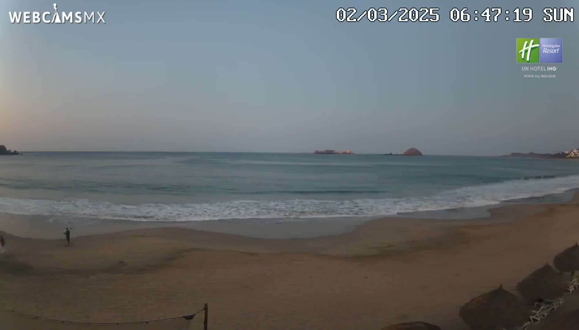 A sandy beach with gentle waves lapping the shore under a clear, pale sky, with a few people visible in the distance.