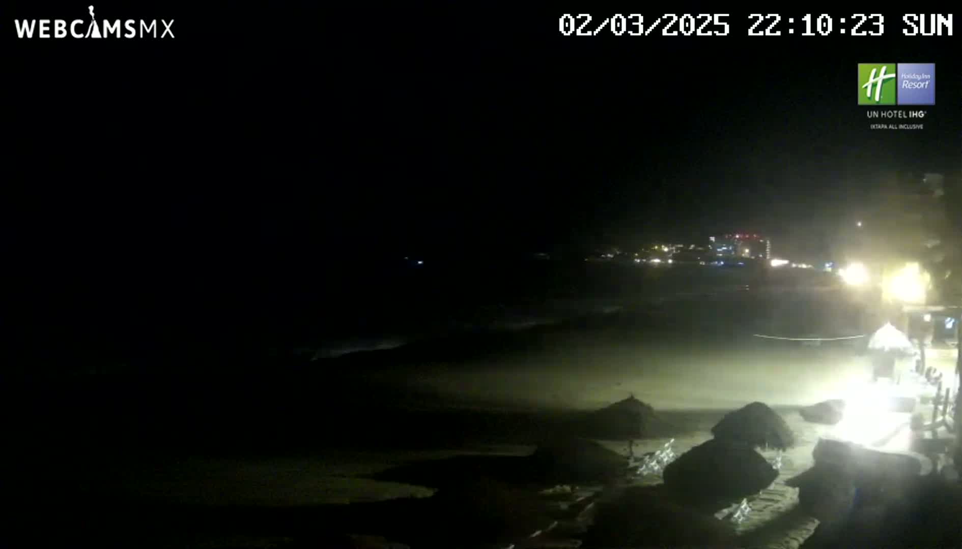 A dark beach at night is partially lit by artificial lights, showing some beach umbrellas and a distant cityscape.
