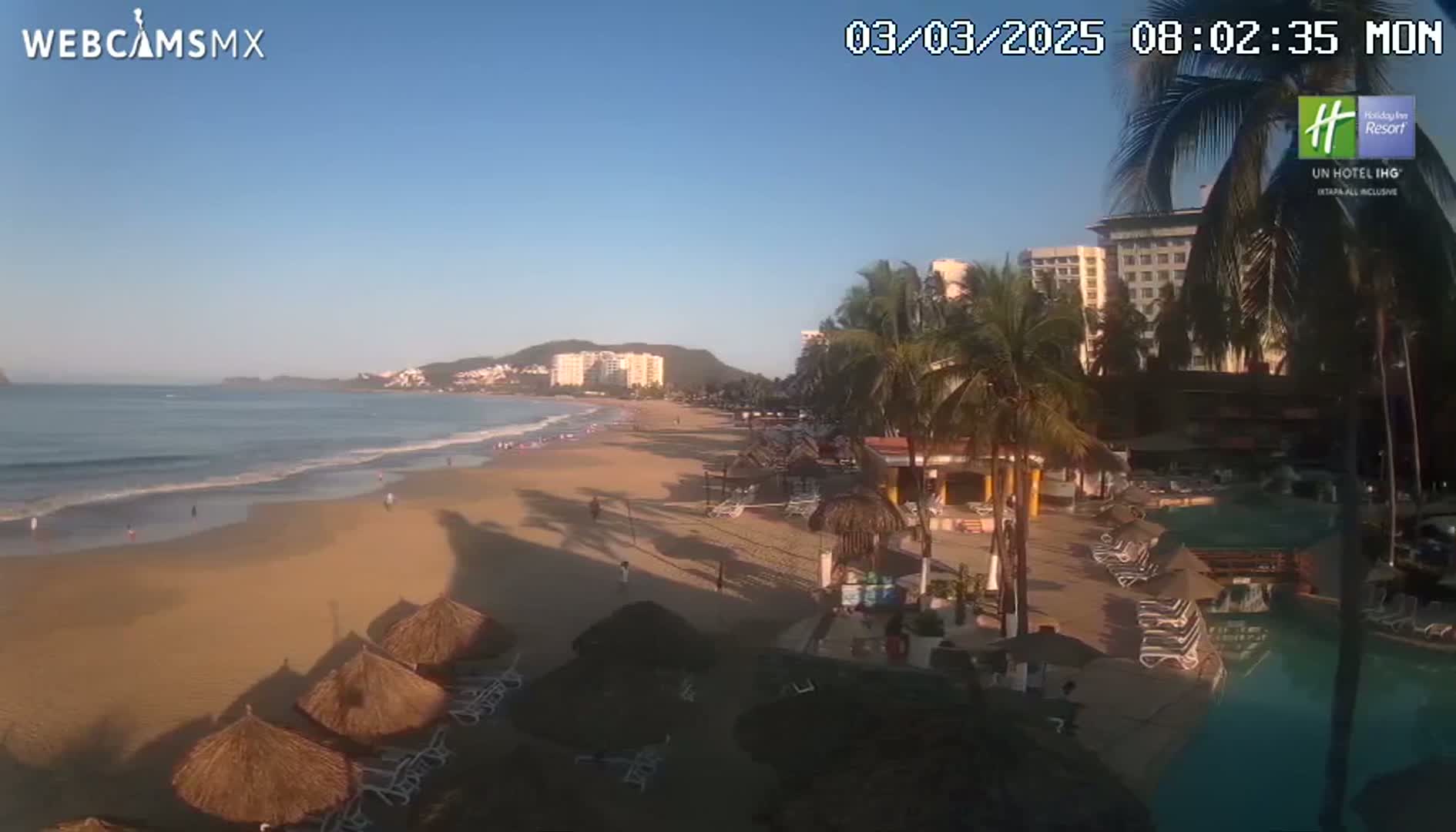 A sandy beach with calm ocean waves is seen from an elevated viewpoint, showing beachfront hotels, palm trees, beach umbrellas, and lounge chairs under a clear, sunny sky.
