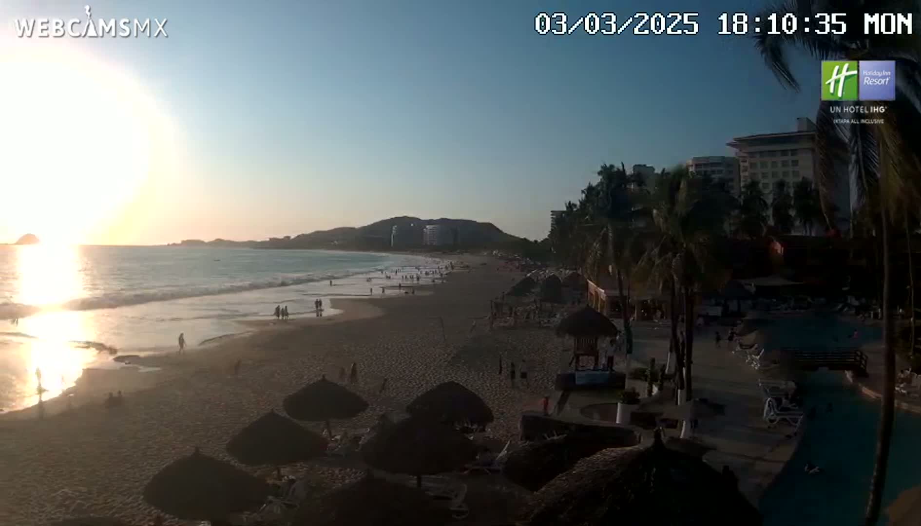 A beach at sunset with calm waves, palm trees, and a resort in the background.