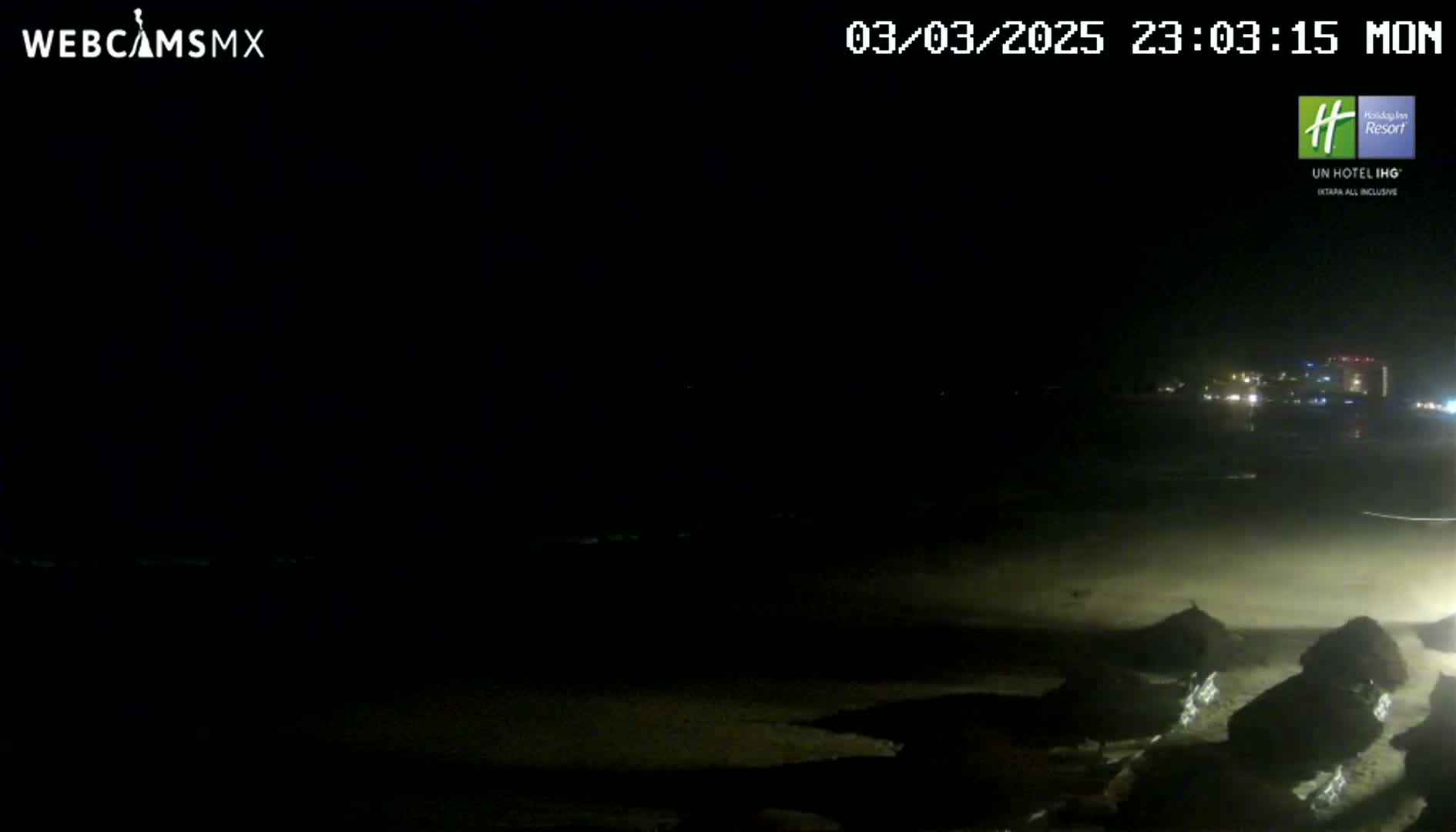 A dark, nighttime beach scene shows a mostly empty sandy shore with some dark objects near the water's edge, under a clear, starless sky.