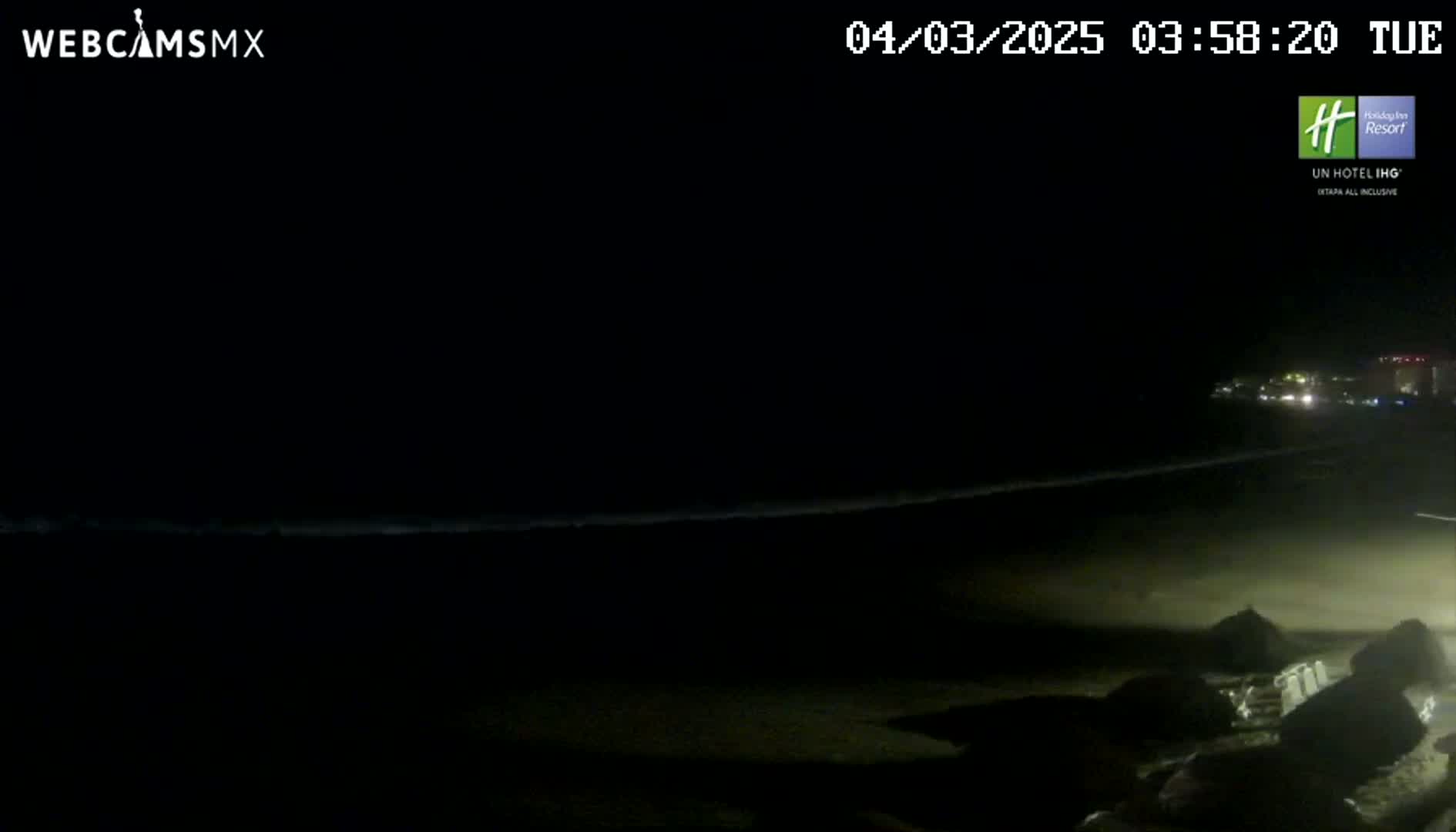 A dark, nighttime beach scene shows a dimly lit shoreline with gentle waves lapping at the sand, under a clear, starless sky.