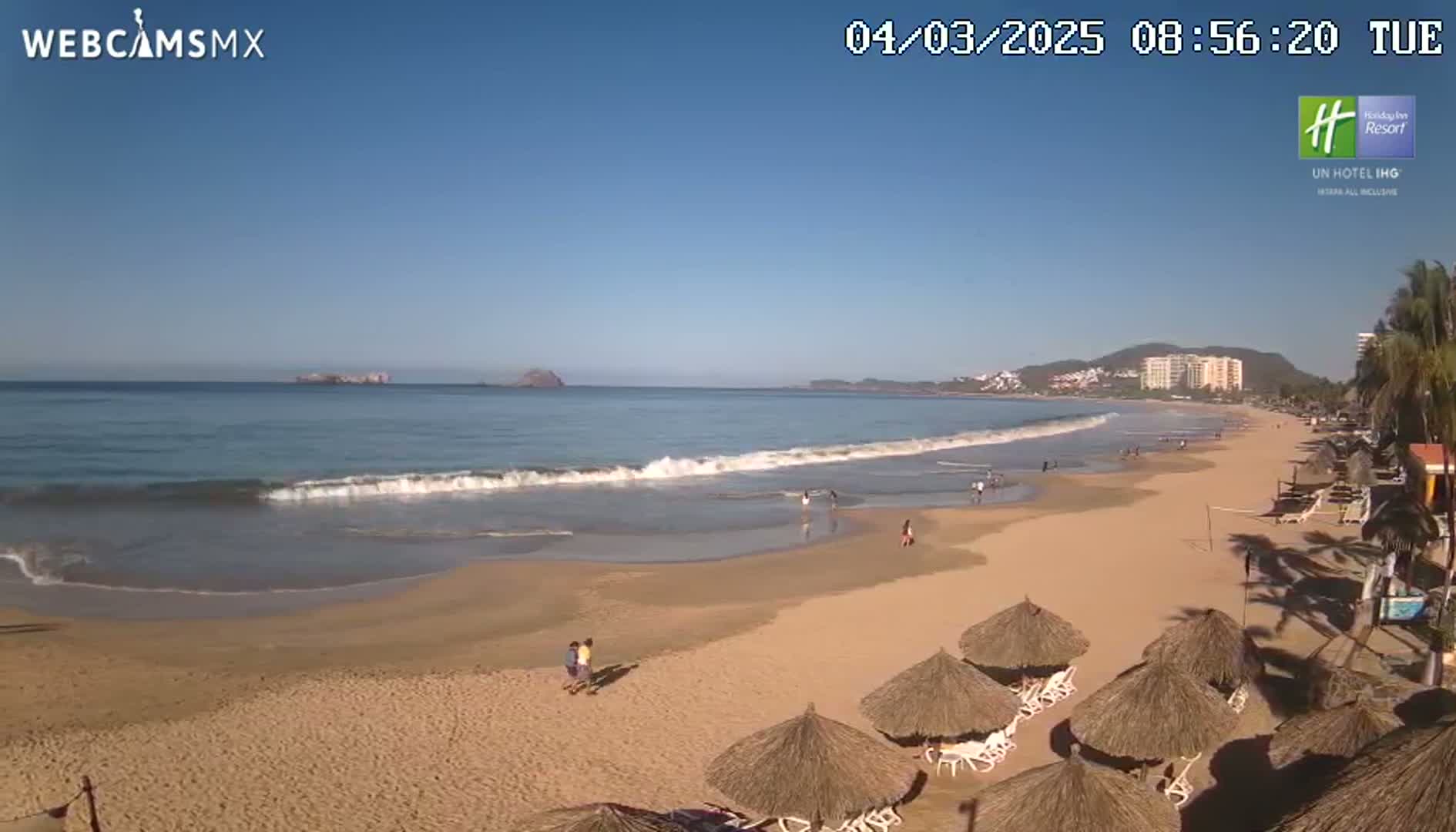 A sandy beach with calm waves under a clear blue sky, lined with thatched umbrellas and beach chairs, and a distant coastline visible.