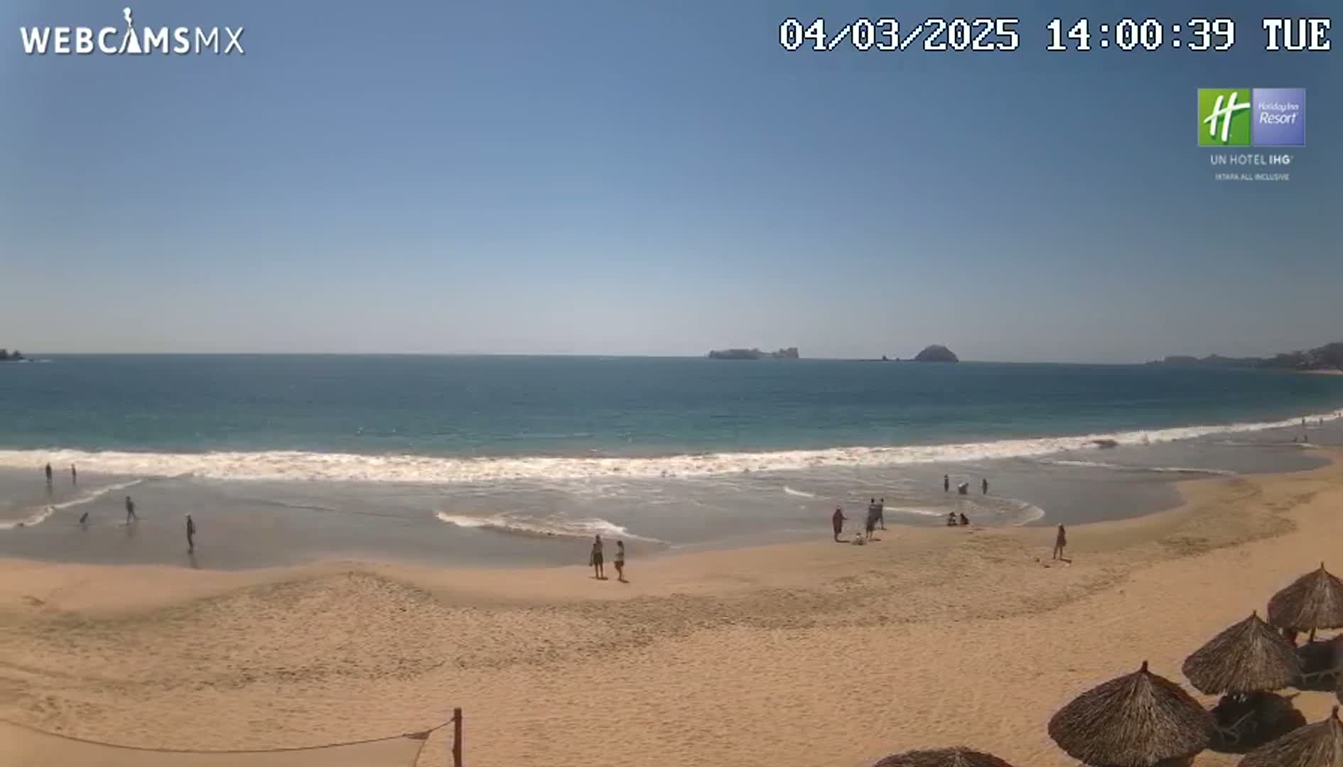 A sandy beach with a few people on it and calm waves rolling onto the shore under a clear blue sky.