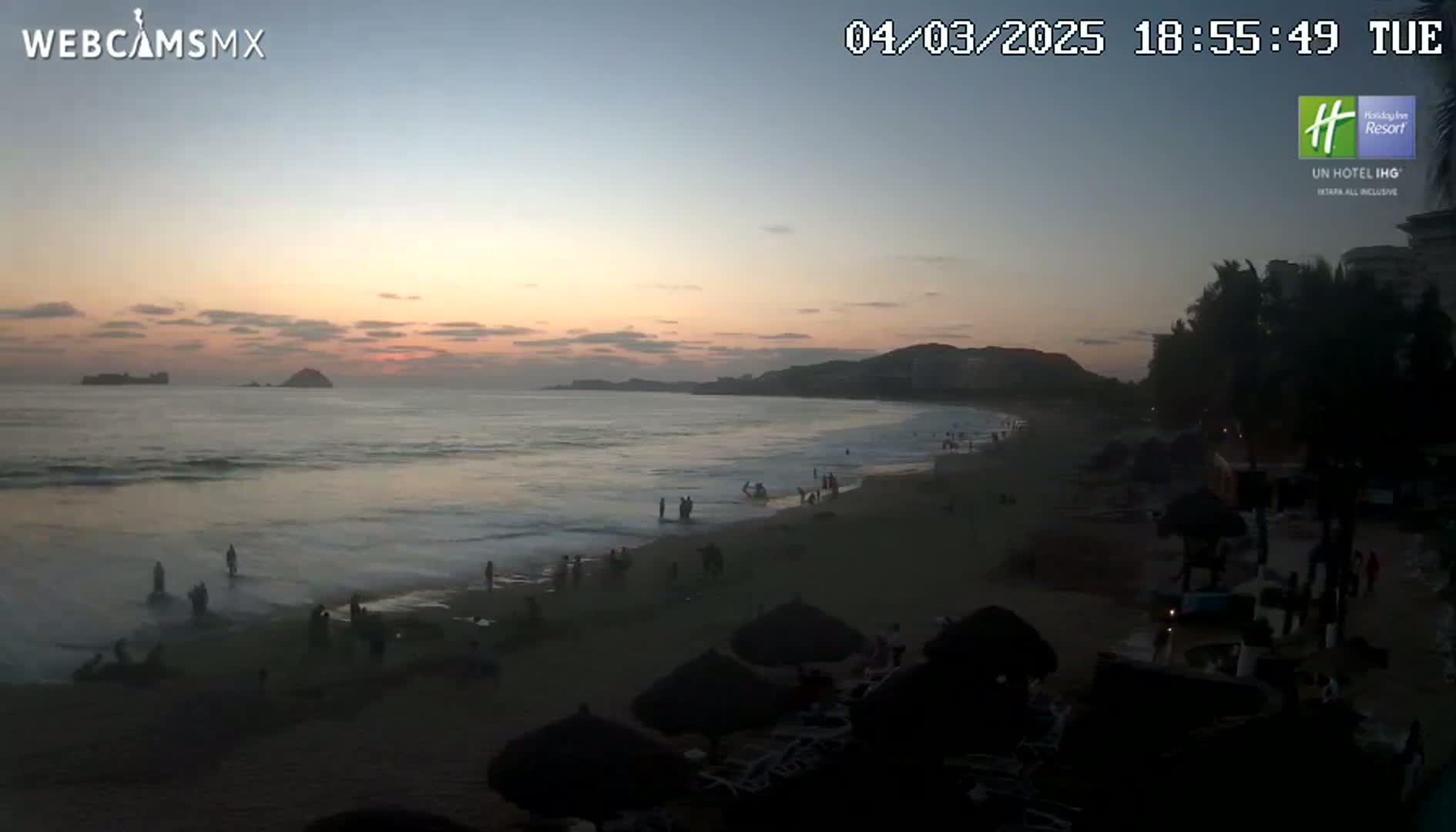 A beach scene at sunset shows a calm ocean with people on the sand, palm trees lining the shore, and hills in the distance.