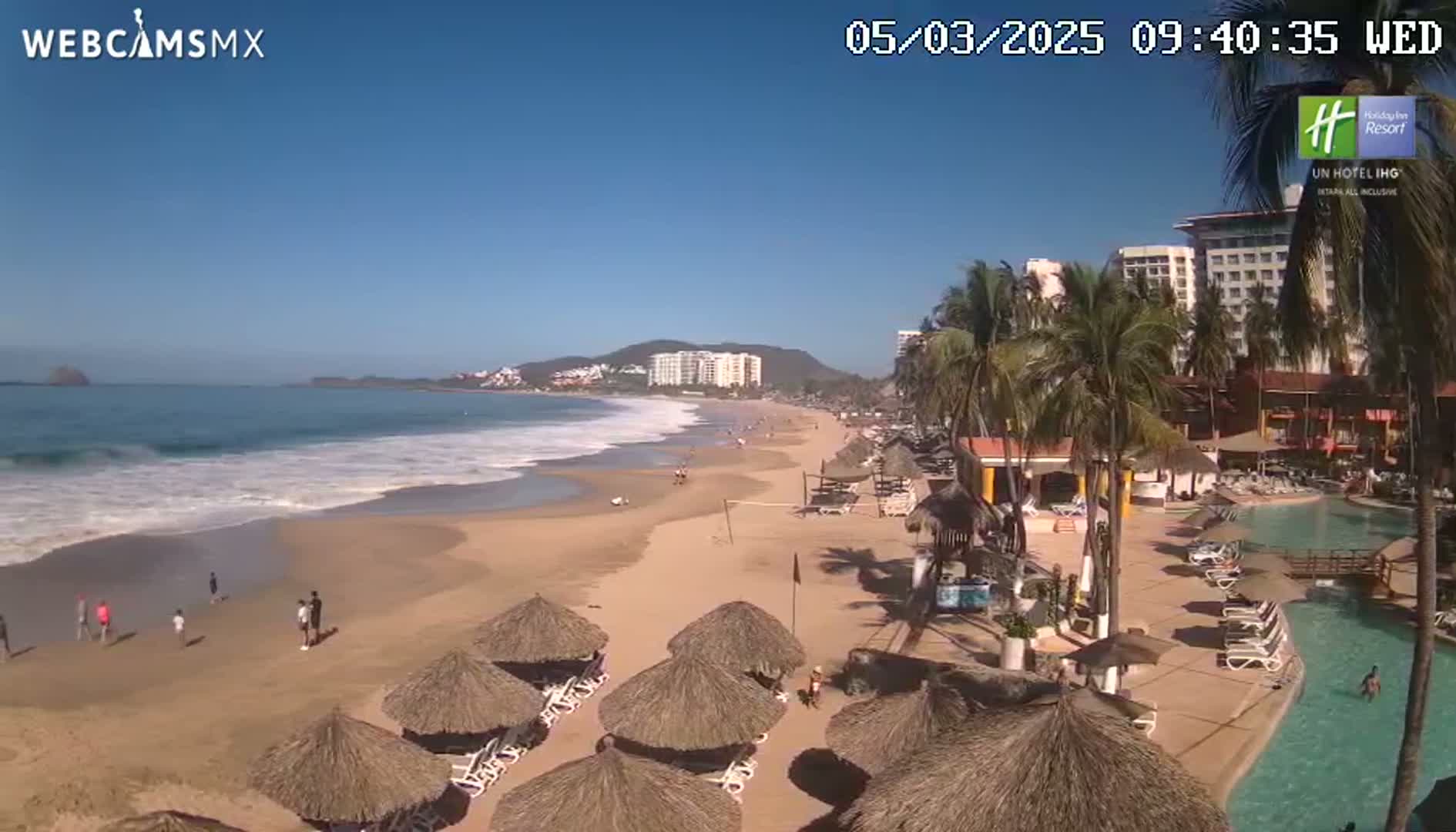 A sandy beach with calm waves, palm trees, beach umbrellas, and a resort pool is seen on a sunny day.