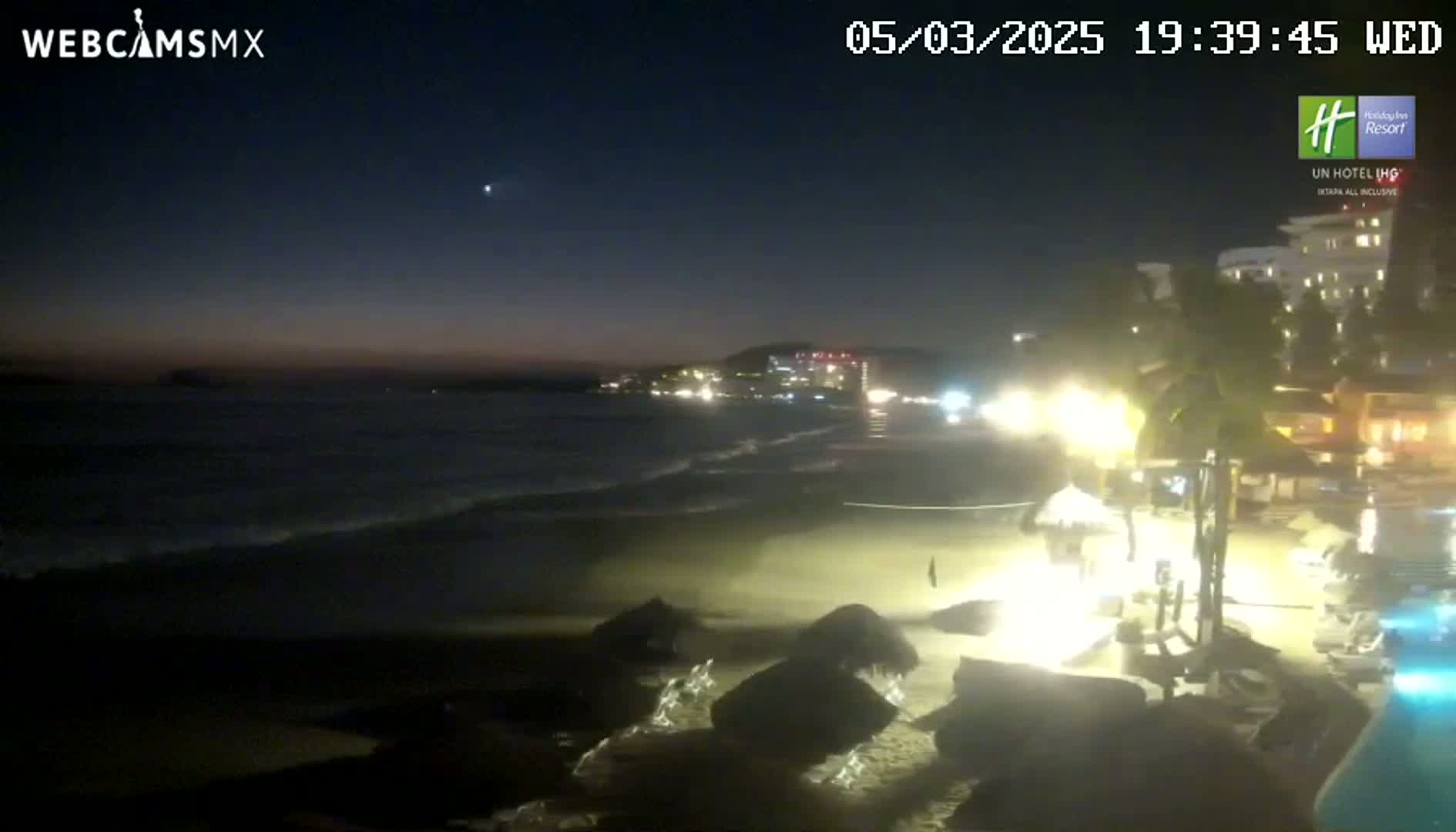 A dark, nighttime beach scene shows gently rolling waves lapping the shore near a brightly lit resort, under a mostly clear sky.