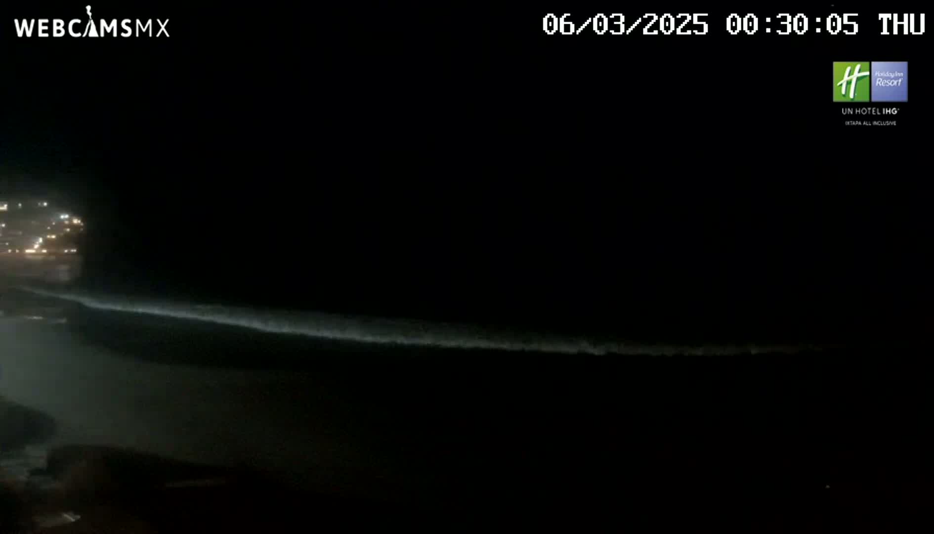 A nighttime view of a dark ocean with a faint line of waves breaking near a dimly lit shoreline.
