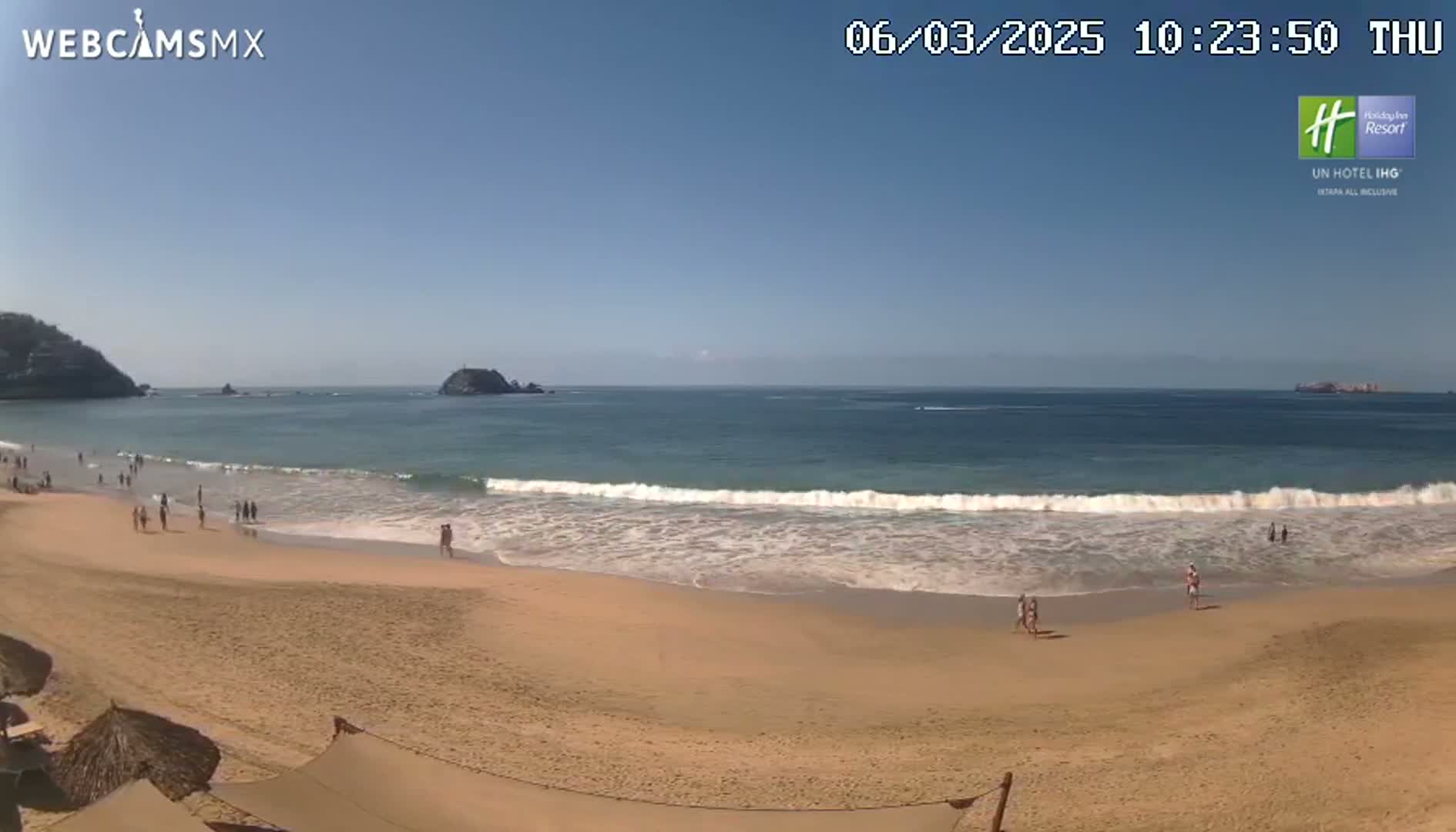A sandy beach with a few people on it is lapped by gentle waves under a clear blue sky.