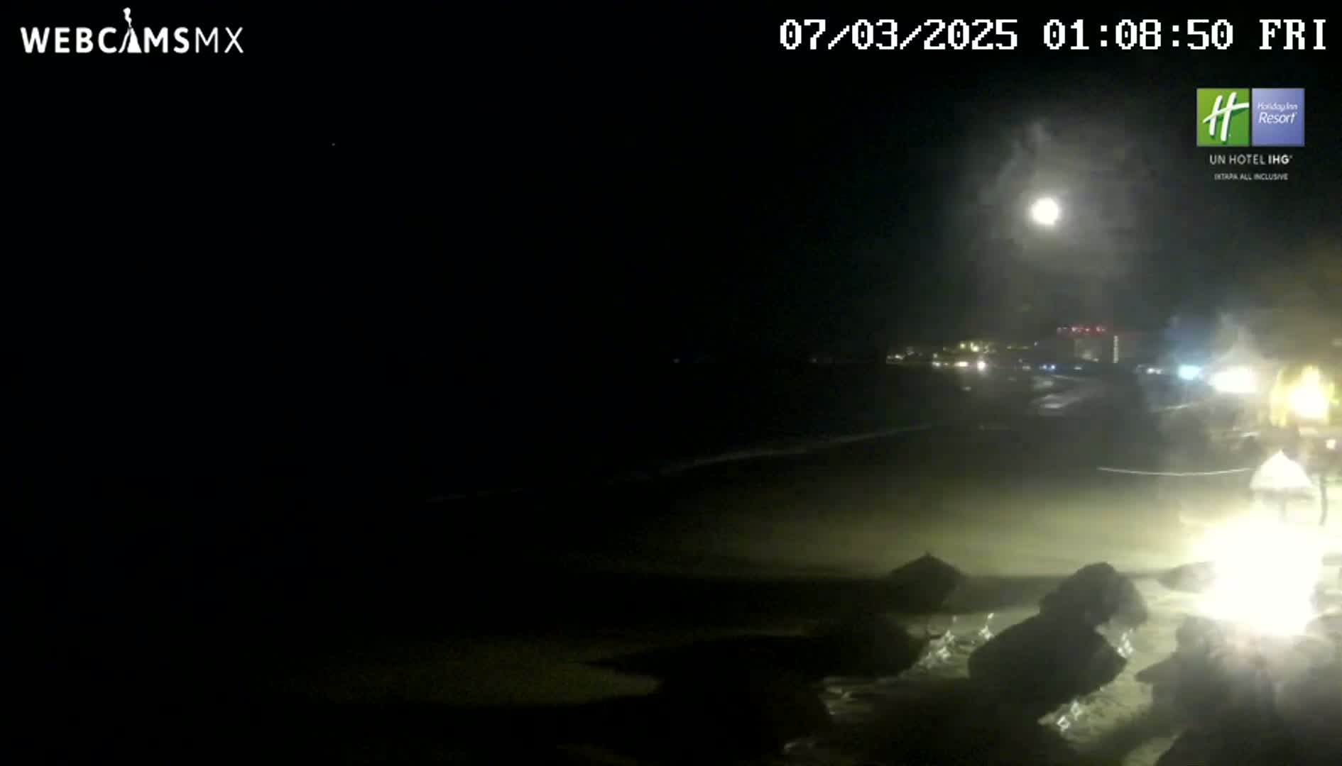 A nighttime beach scene shows a bright moon in the sky, dark sand, and some rocks near the water's edge, with faint lights visible in the distance.
