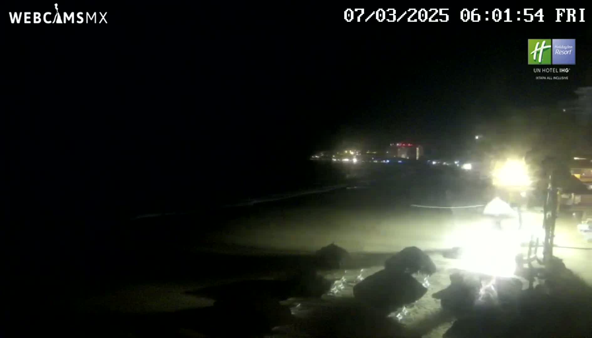 A dark, nighttime beach scene shows a dimly lit shoreline with rocks in the foreground and distant lights from buildings across the water.