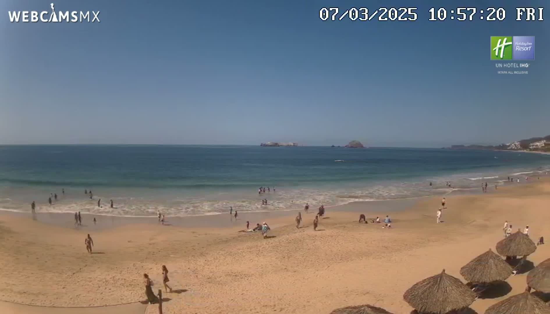 A sandy beach with calm ocean waves and numerous people enjoying the sunny day.