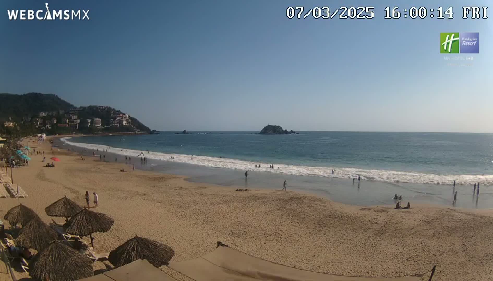 A sandy beach with calm ocean waves stretches out under a clear blue sky, populated by scattered beachgoers and shaded by thatched umbrellas.