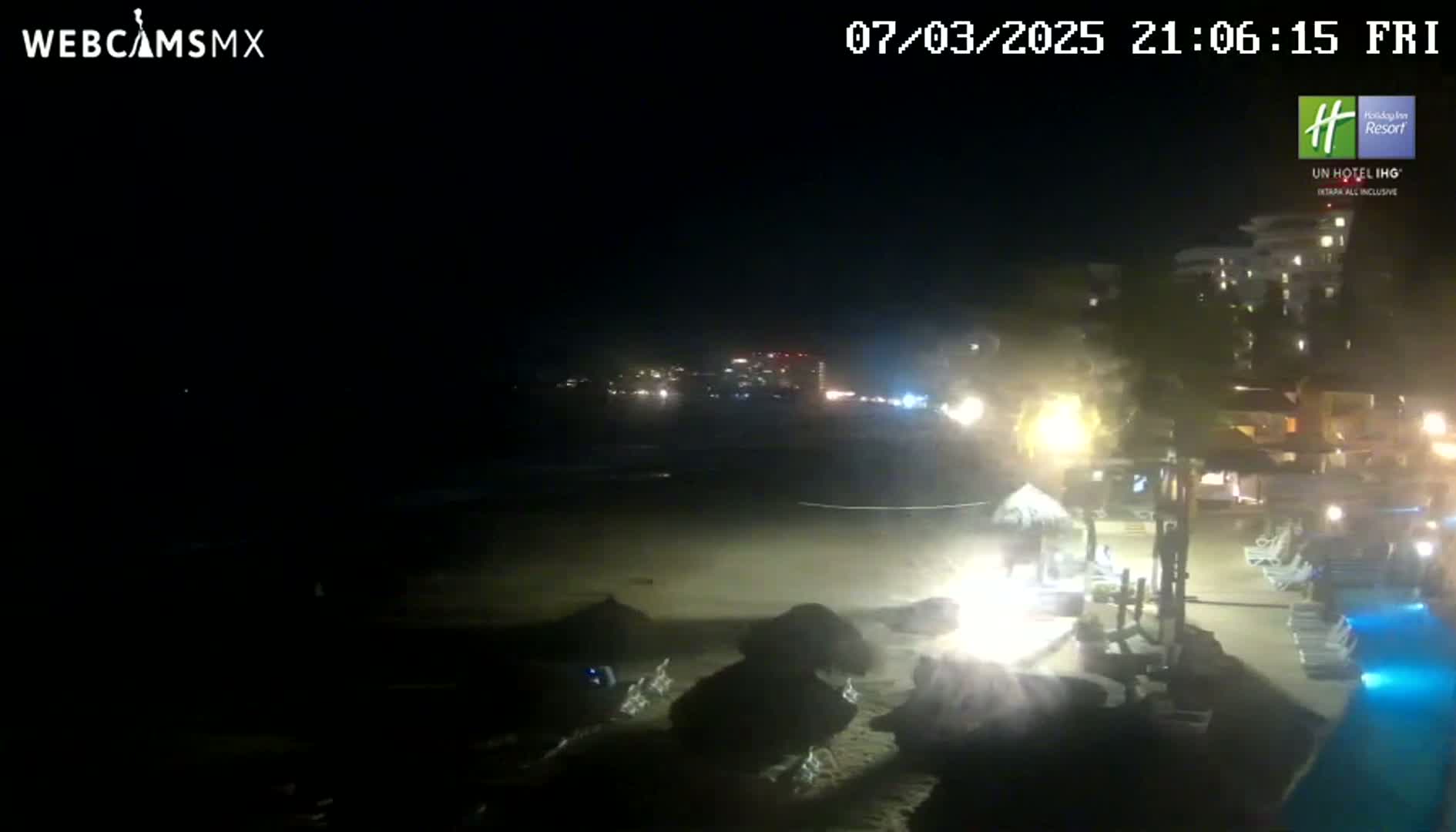 A nighttime beach scene shows a dark ocean with waves lapping the shore,  illuminated buildings in the background, and dimly lit beach furniture near a pool.