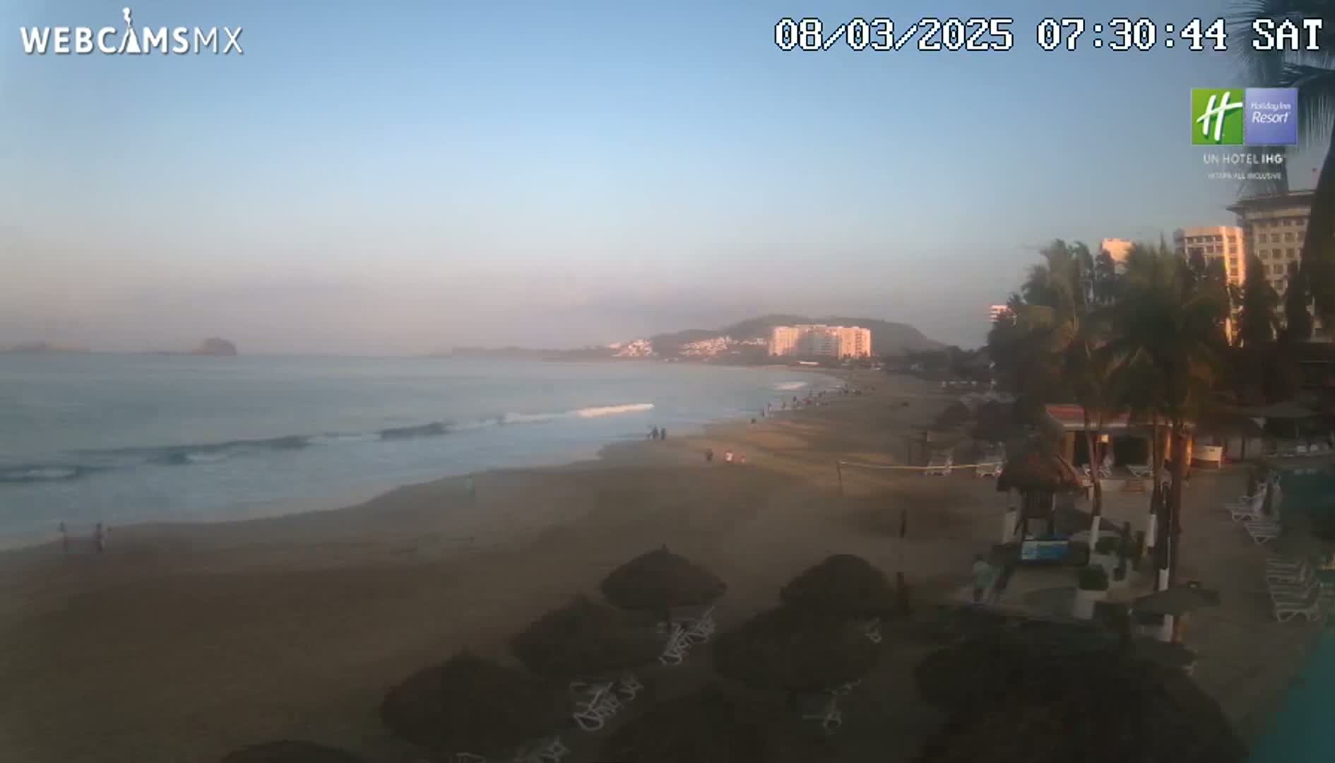 A sandy beach with gentle waves under a clear, dawn sky, showing a beachfront with buildings in the distance.