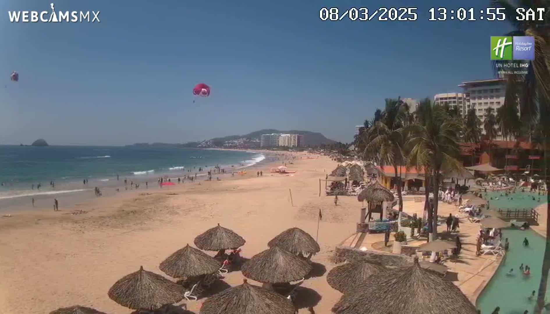 A sandy beach with calm ocean waves, thatched umbrellas, and palm trees, under a sunny sky, features parasailers and people enjoying the water and shore.