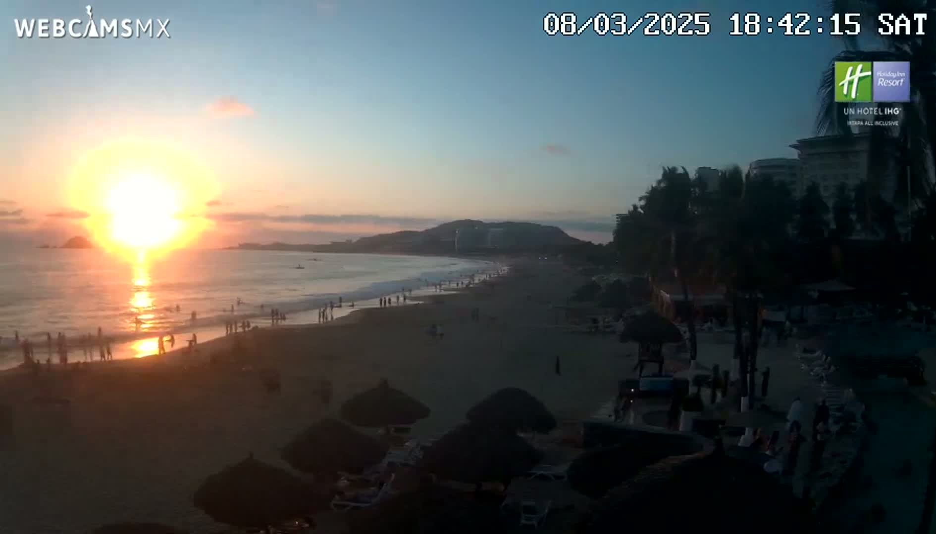 A sunset over a beach with people enjoying the sand and ocean, under a clear sky.