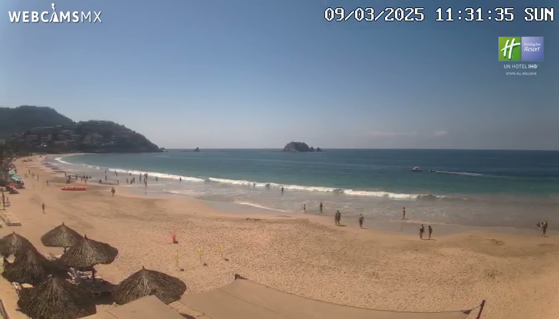 A sandy beach with calm ocean waves under a clear blue sky, dotted with people and thatched umbrellas.