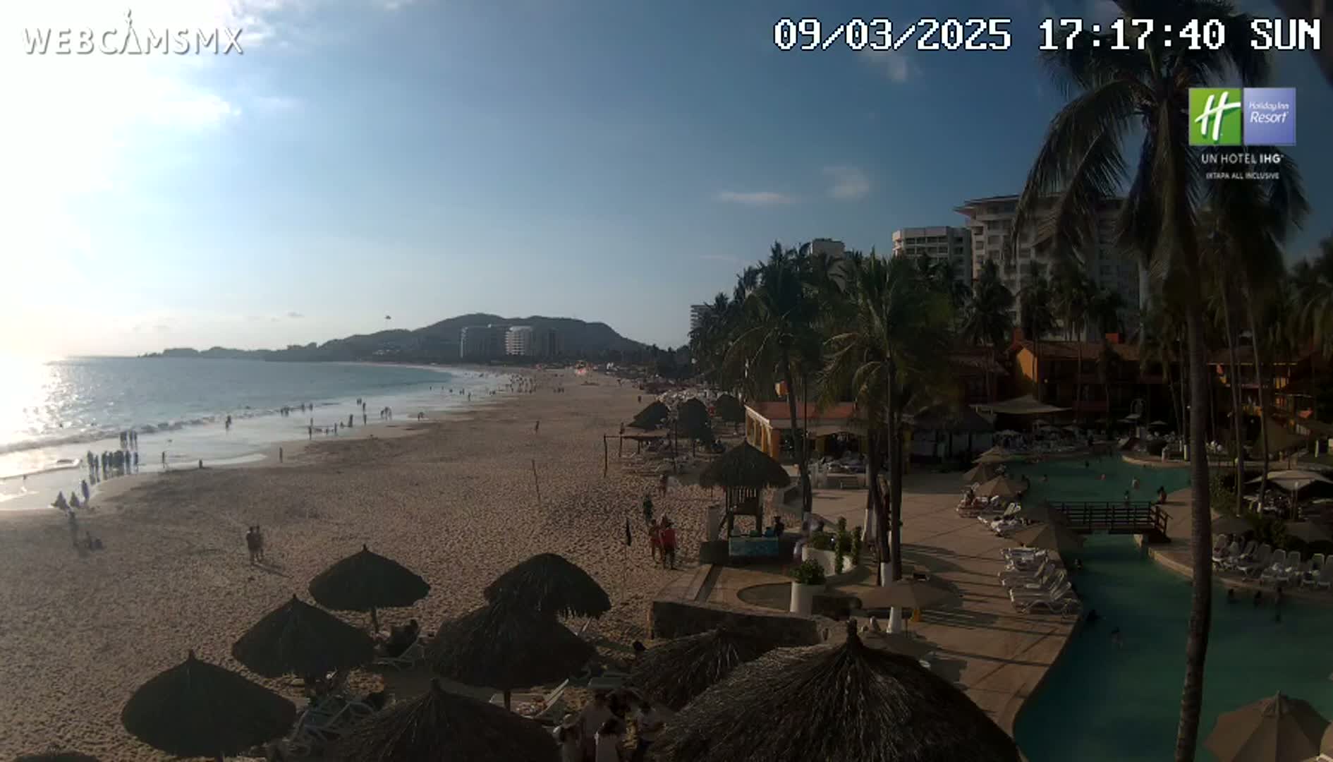 A sunny beach scene shows a sandy shore with scattered beachgoers, palm trees, resort buildings, and a swimming pool, all under a clear sky.