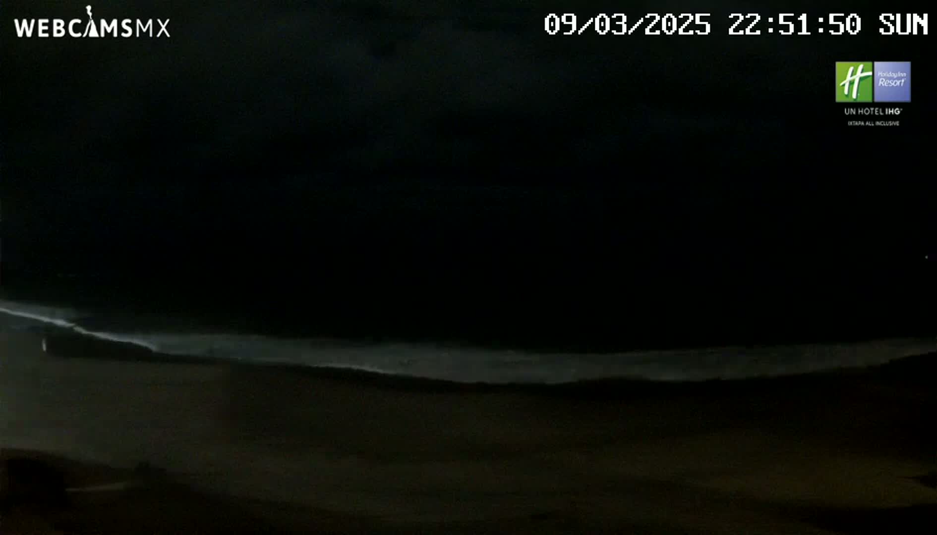 A dark nighttime beach scene shows a faint line of waves breaking on the shore under a cloudy sky.