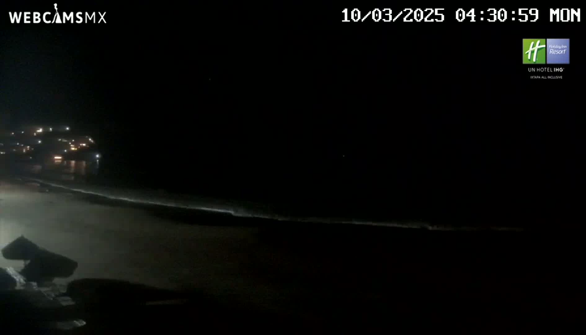 A dark, nighttime beach scene shows a dimly lit shoreline with a building in the distance under a clear, starlit sky.