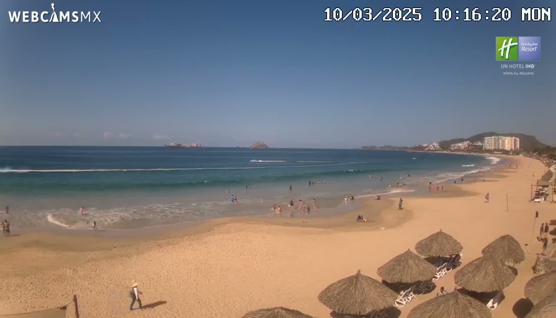 A sandy beach with calm ocean waves and scattered people under thatched umbrellas on a sunny day.