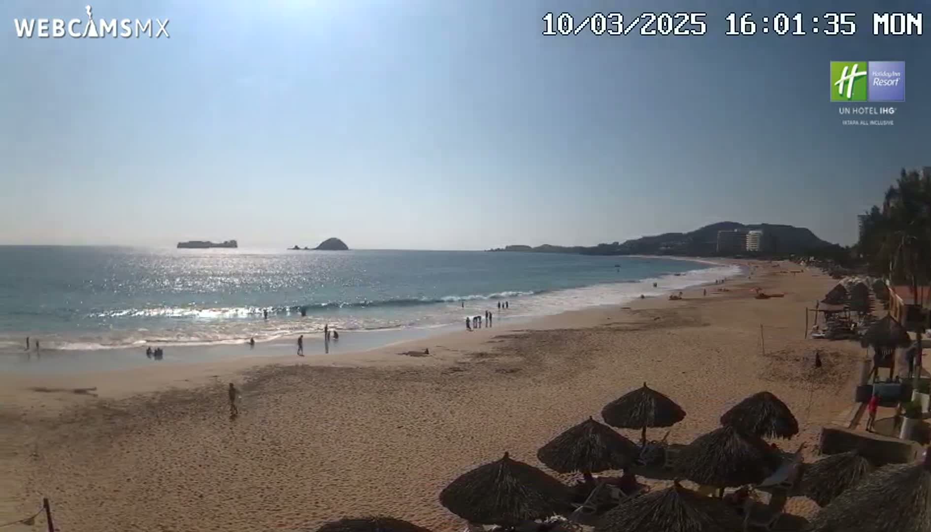 A sandy beach with calm ocean waves under a clear, sunny sky is dotted with people and thatched umbrellas.