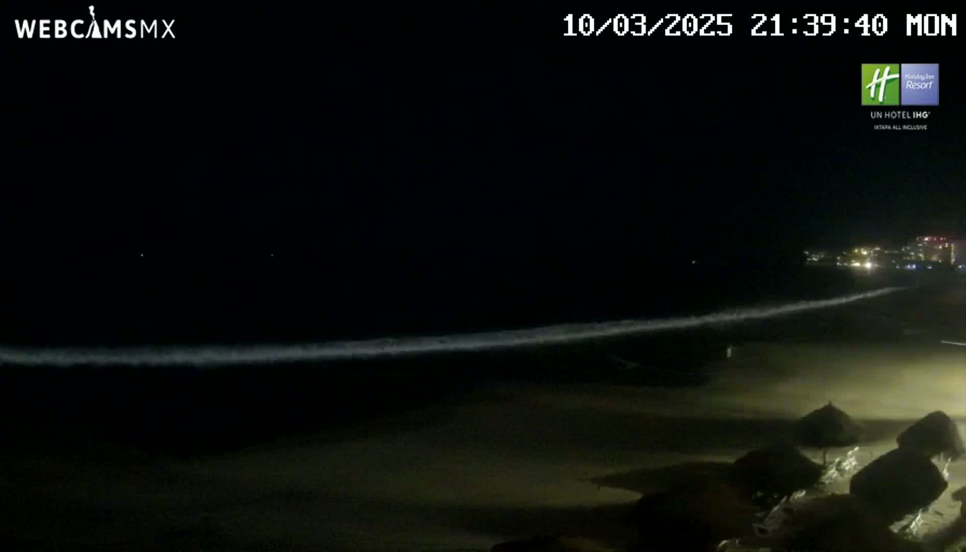 A dark beach at night shows gently lapping waves under a clear, moonlit sky; several dark beach umbrellas are visible in the foreground.