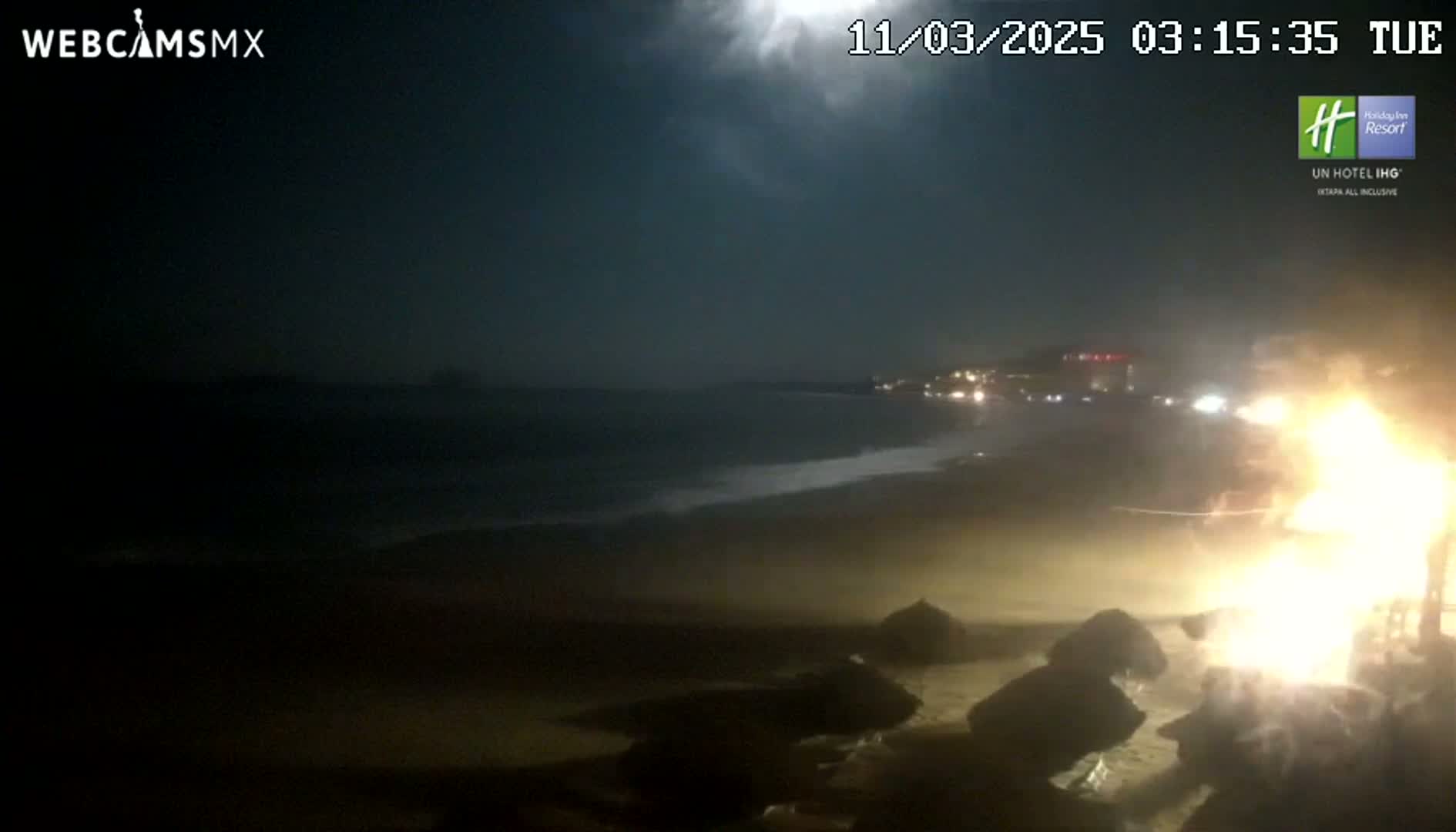 A nighttime beach scene shows a dark ocean with gentle waves under a mostly cloudy sky, with a bright fire burning near the shore.