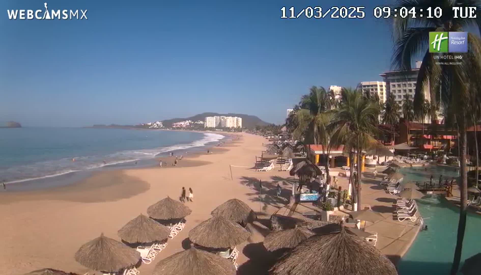 A sandy beach with calm ocean waves, numerous thatch-roofed umbrellas, a resort pool, and palm trees under a clear blue sky.