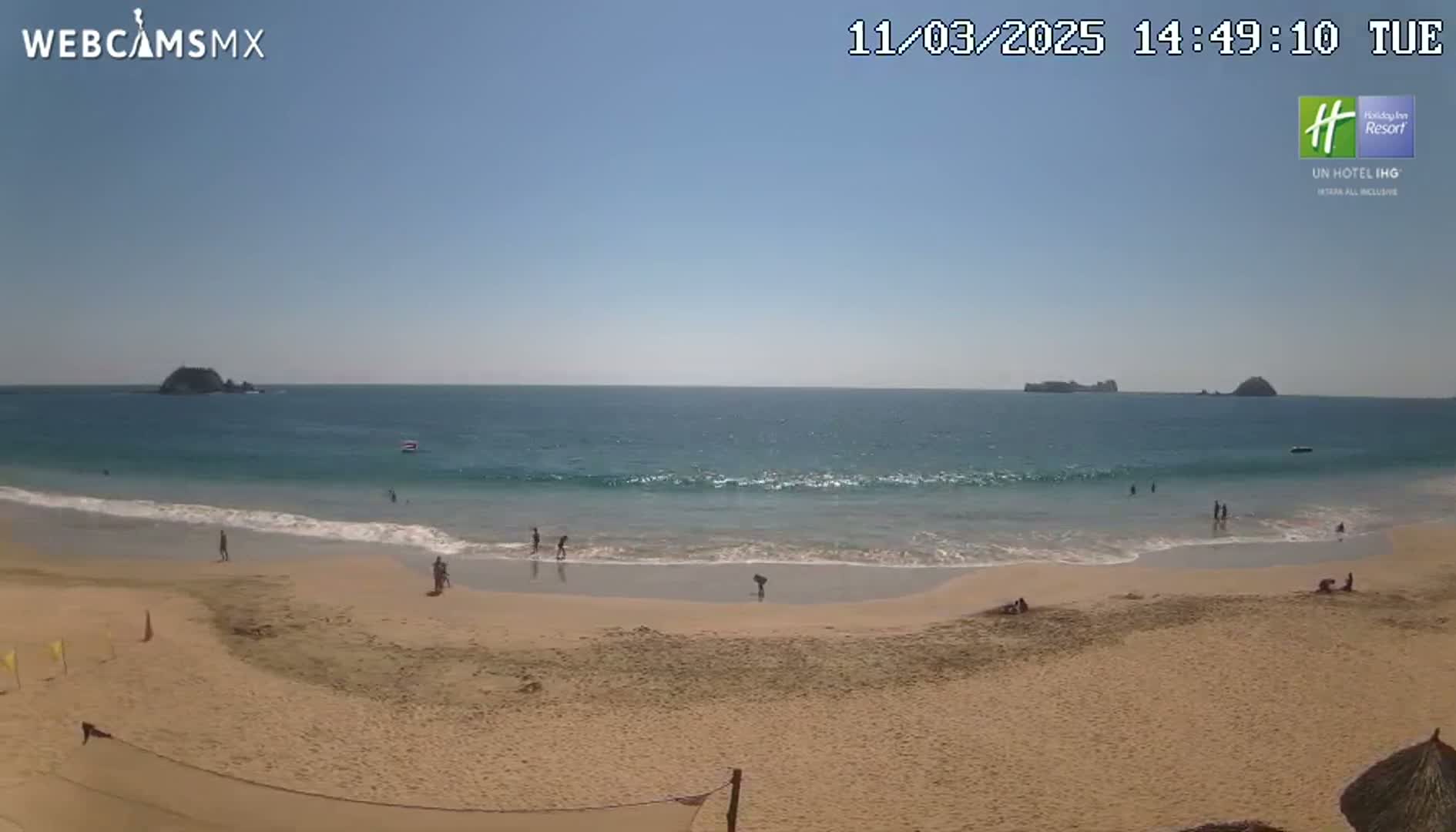 A sandy beach with a few people on it slopes down to a calm ocean with small waves under a clear blue sky.