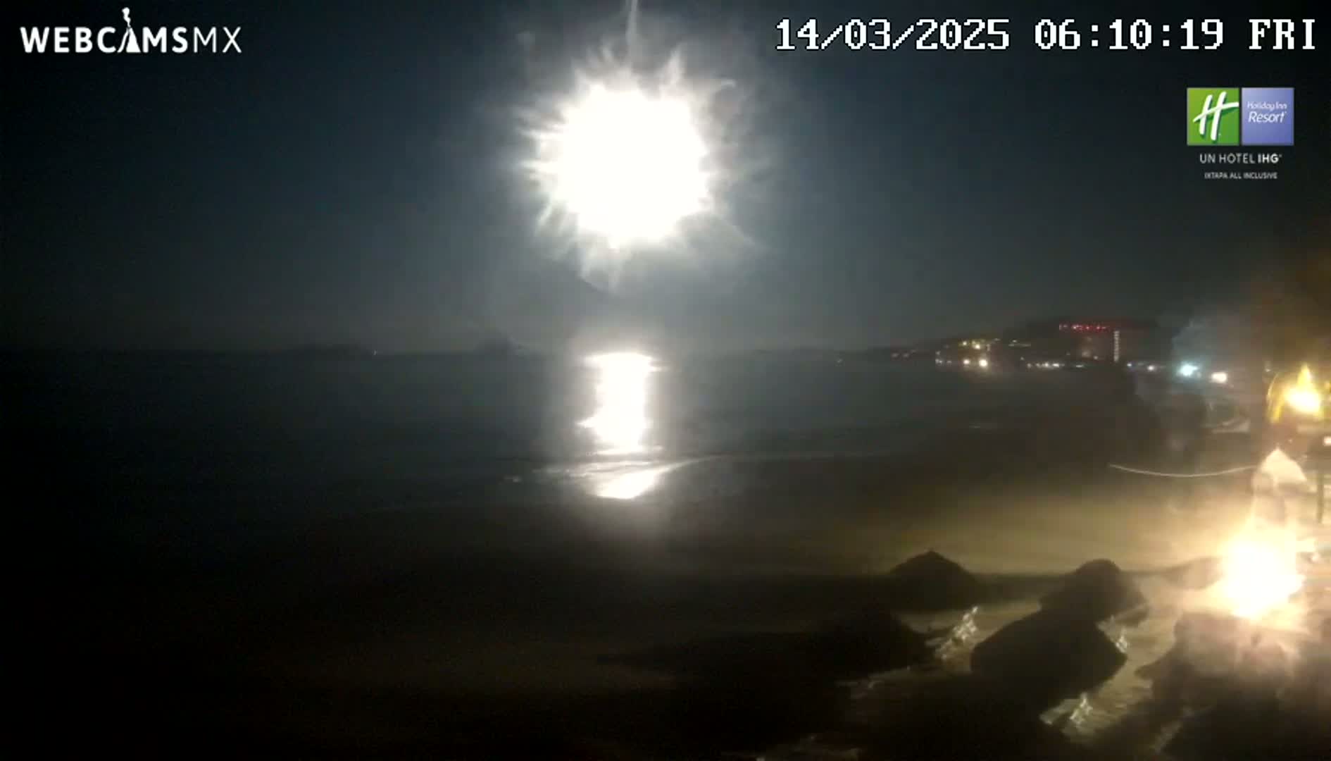 A bright moon shines over a dark beach and ocean at night, illuminating the water and shoreline.
