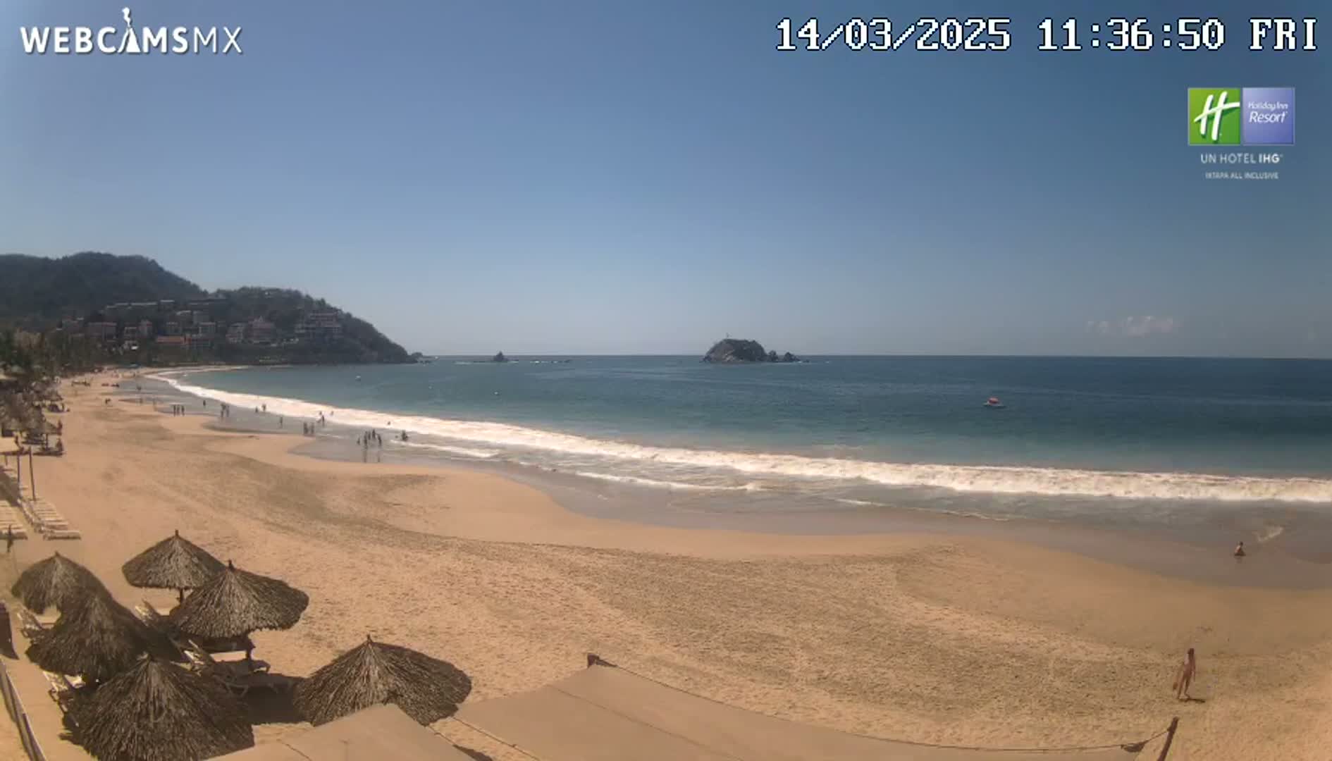 A sandy beach with calm waves, several thatched umbrellas, and a few people are visible under a clear, sunny sky, with hills visible in the distance.