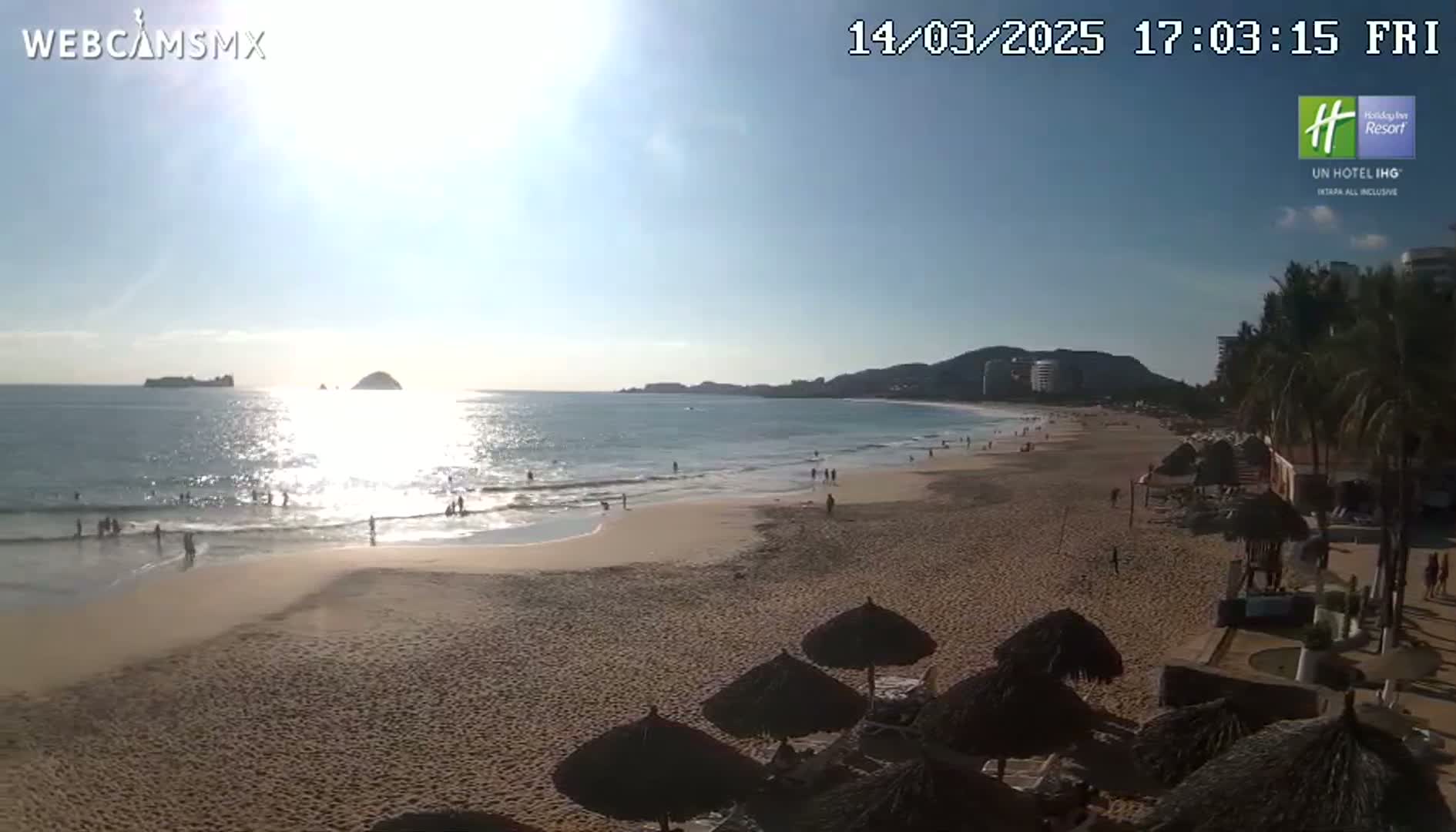 A sunny beach scene shows a wide sandy shore with calm ocean waves, a few people enjoying the water, and several thatched umbrellas near the buildings along the coastline.